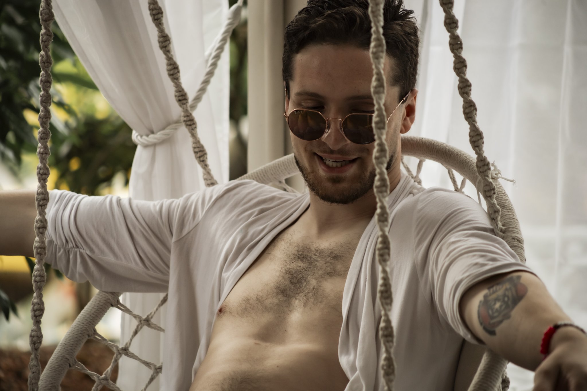 a male wearing sunglasses as he sits outside on a hammock chair with sheer curtains behind him.
