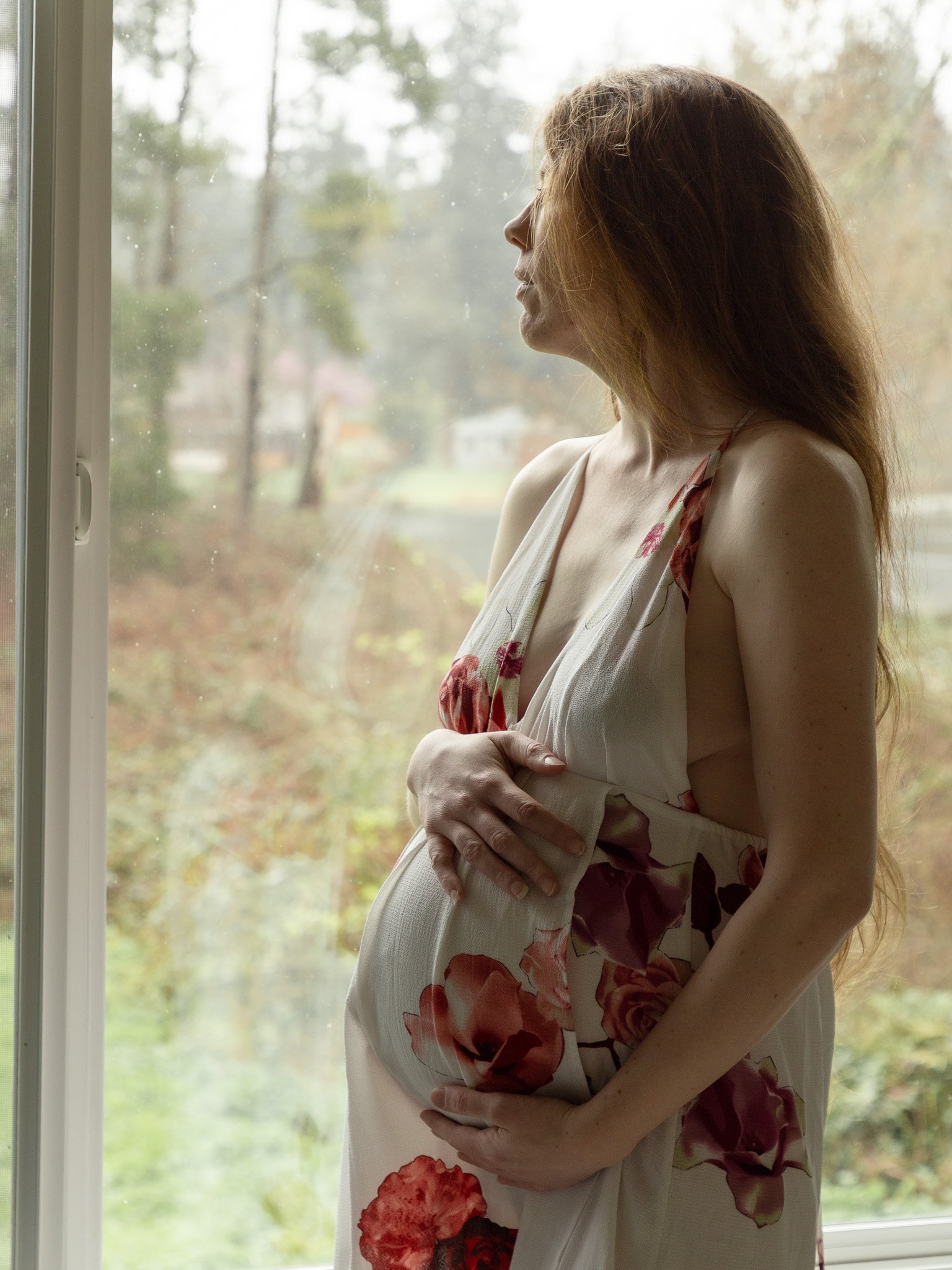 A woman in a floral dress standing by a window, looking outdoors with her hand on her pregnant belly.