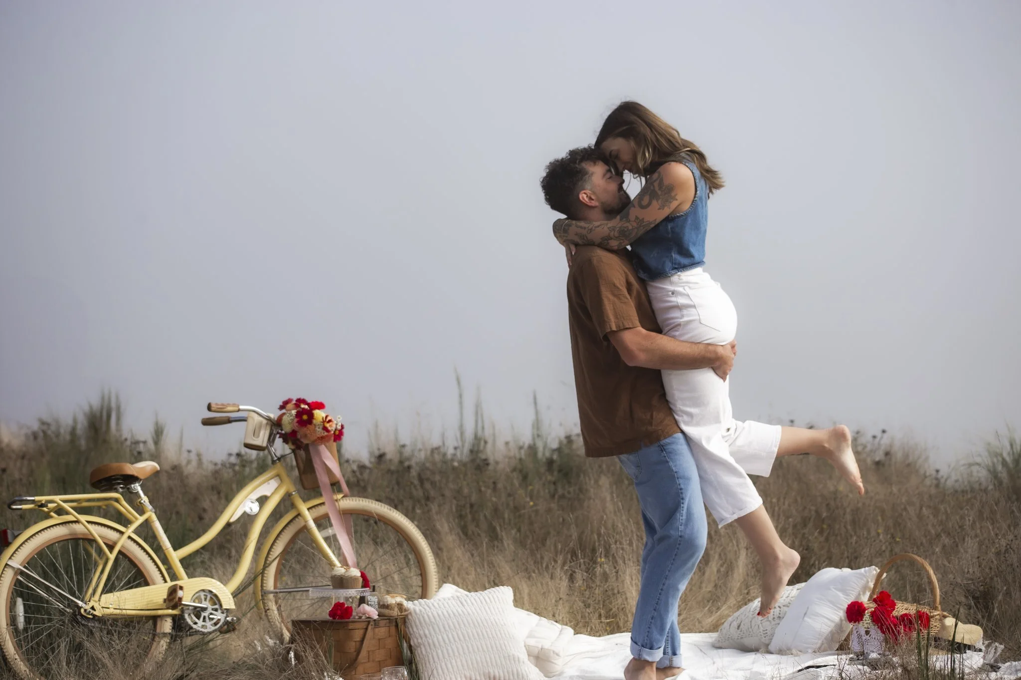 A couple sharing a joyful moment outdoors during a picnic, with the man lifting the woman in the air, and a yellow bicycle with flowers attached to the front in the background.