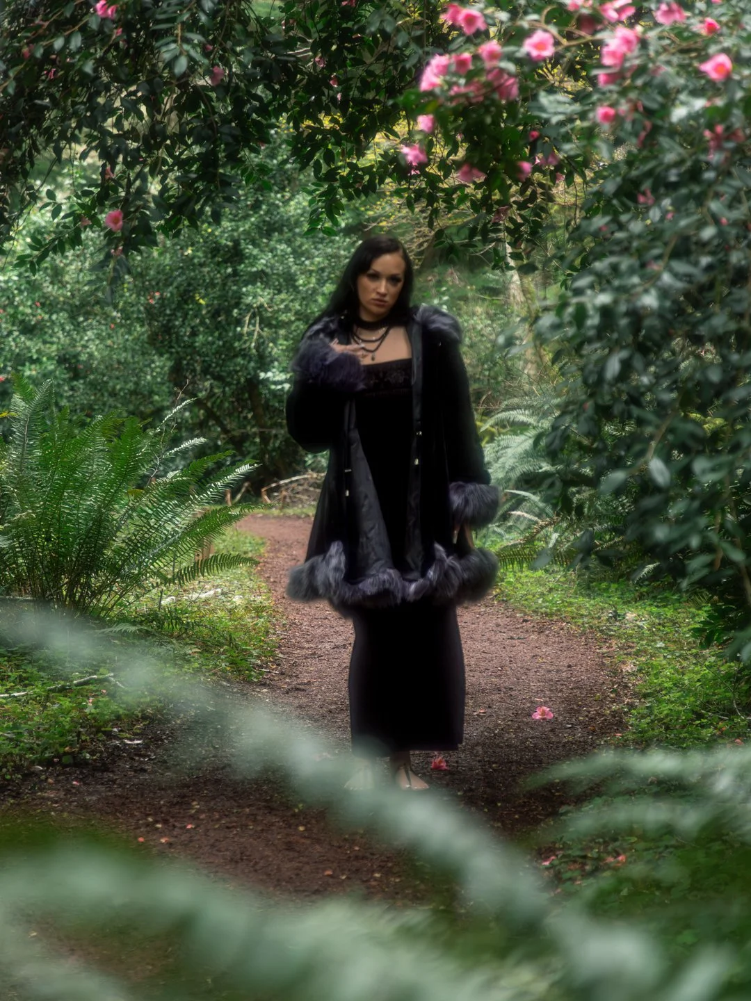 A woman dressed in a black outfit with a coat featuring fur trim standing on a forest path surrounded by greenery and pink flowers.
