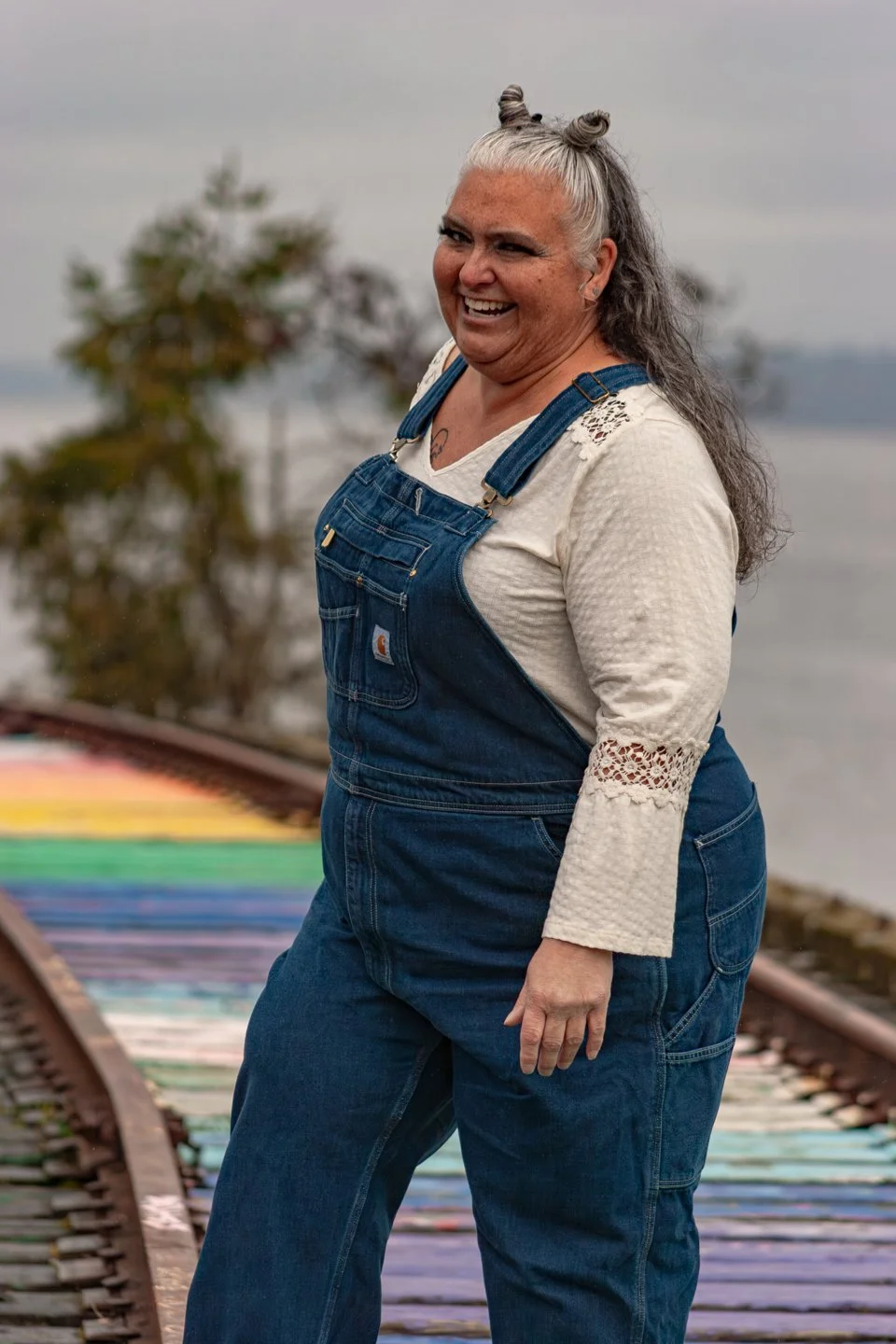 A woman with gray and black hair styled in space buns, wearing a cream long sleeve top with lace details and blue denim overalls, standing outdoors on train tracks painted in rainbow colors, smiling at the camera with trees and a body of water blurred in the background.