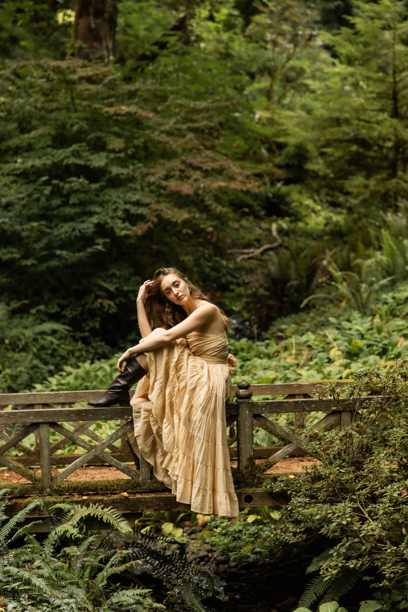 A woman in a flowing beige dress and black knee-high boots sitting on a wooden bridge railing in a lush green forest. She has light brown hair and is touching her hair with one hand while gazing thoughtfully.