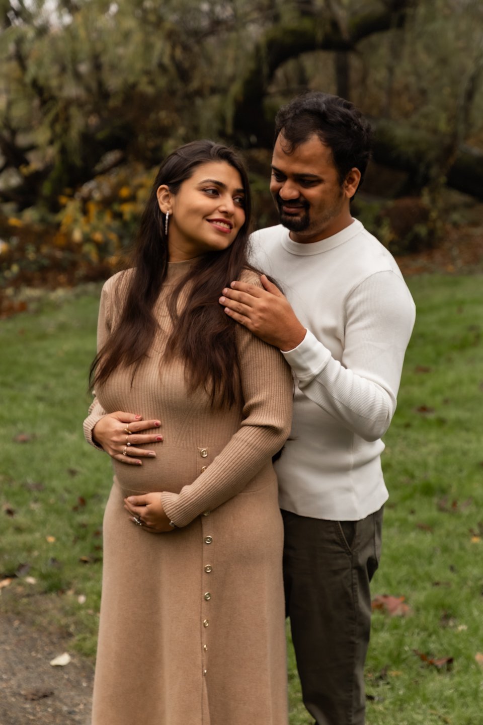A smiling pregnant woman in a beige dress with buttons down the front, standing outdoors on grass with trees and fallen leaves in the background, being embraced by a man in a white long-sleeve shirt.