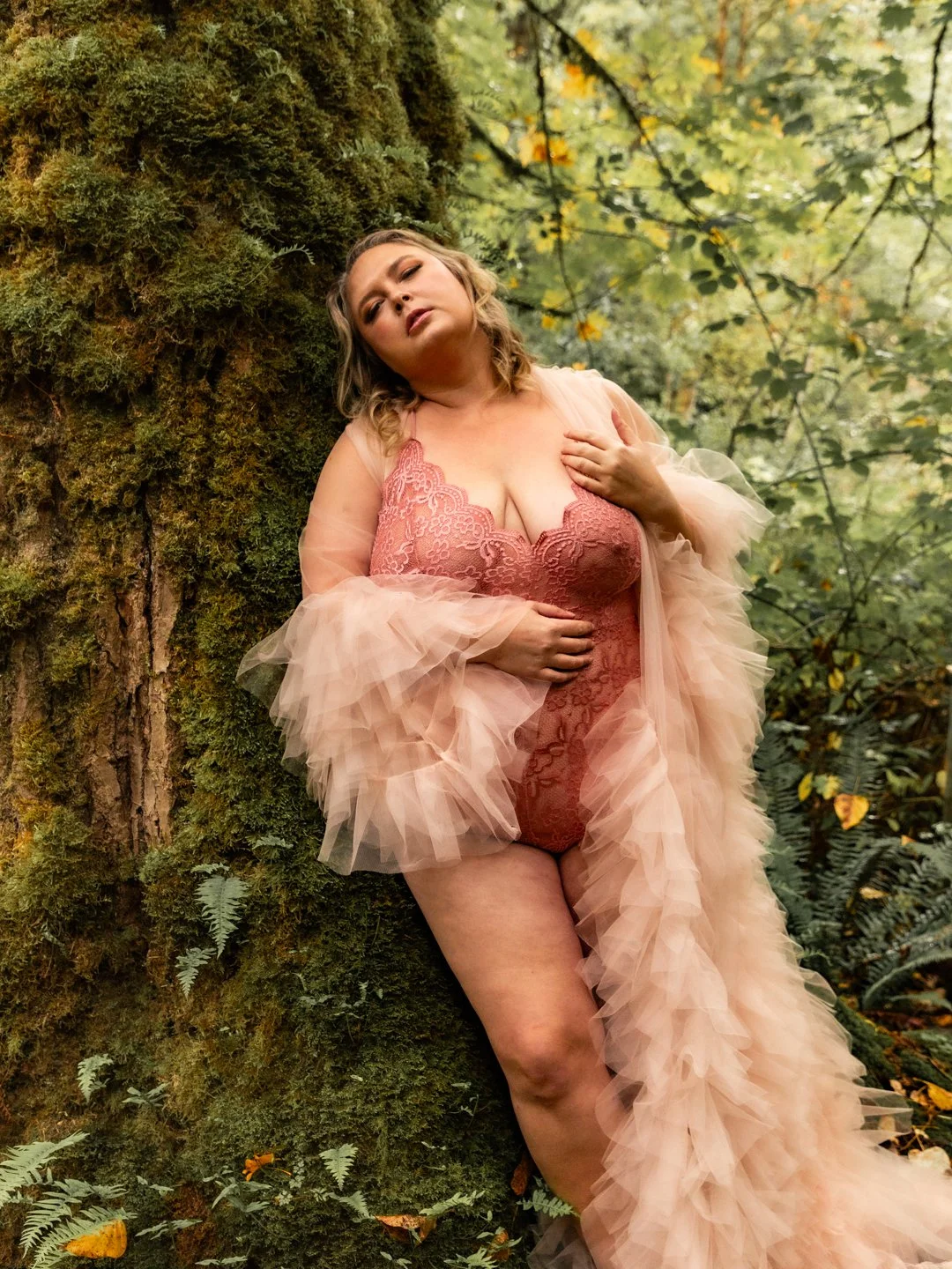 A woman wearing a pink lace bodysuit and a fluffy, sheer pink robe stands outdoors, leaning against a moss-covered tree trunk, with green foliage in the background.