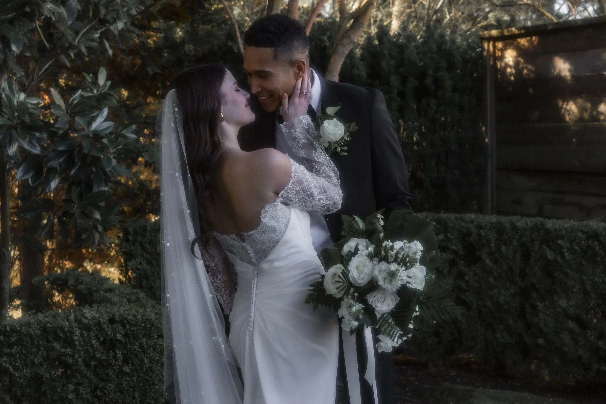 A bride and groom share a tender moment during their wedding, with the bride gently touching the groom's face, set outdoors amidst trees and greenery at sunset.