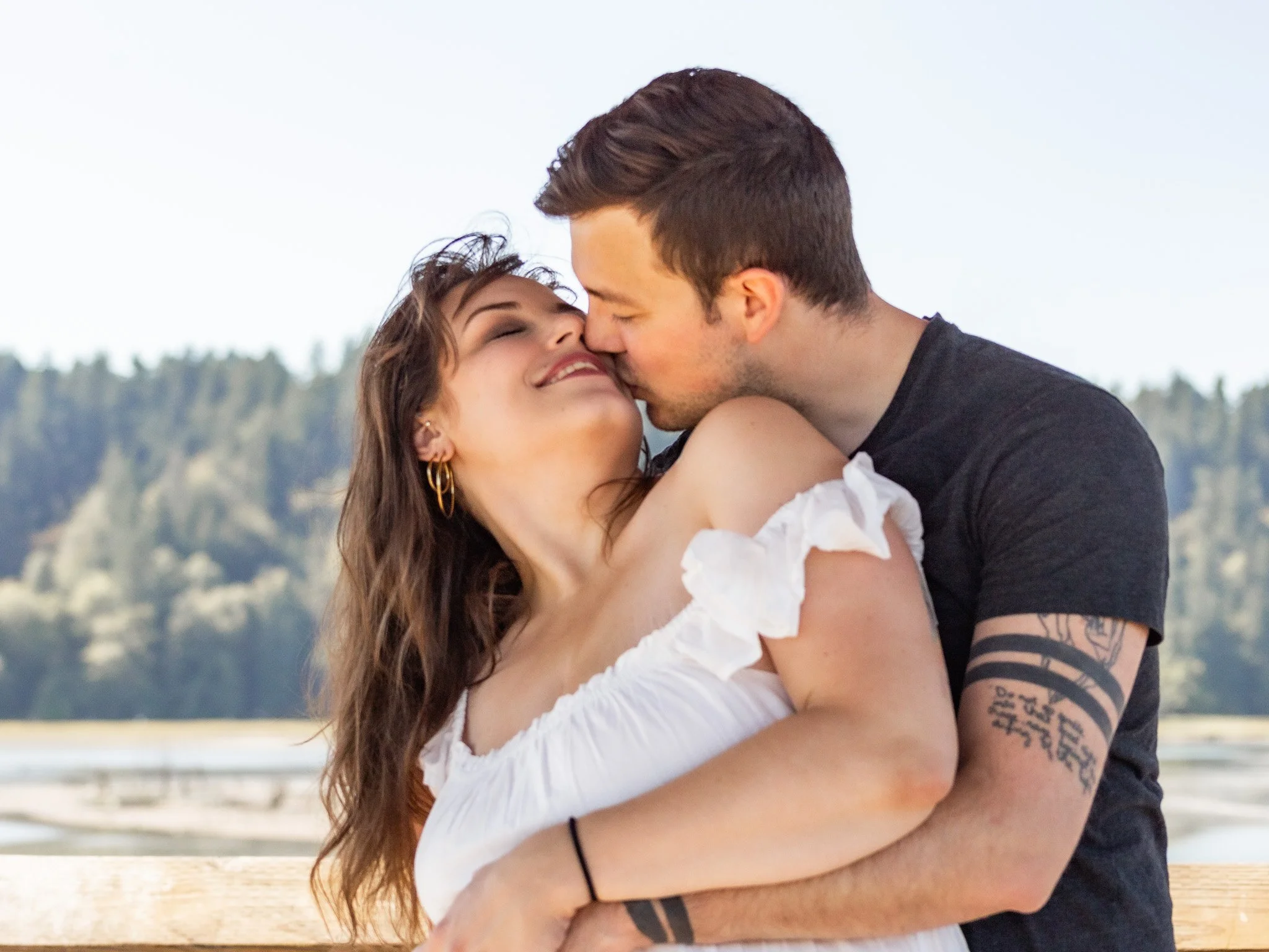 A couple hugging and kissing outdoors with trees and a cloudy sky in the background.