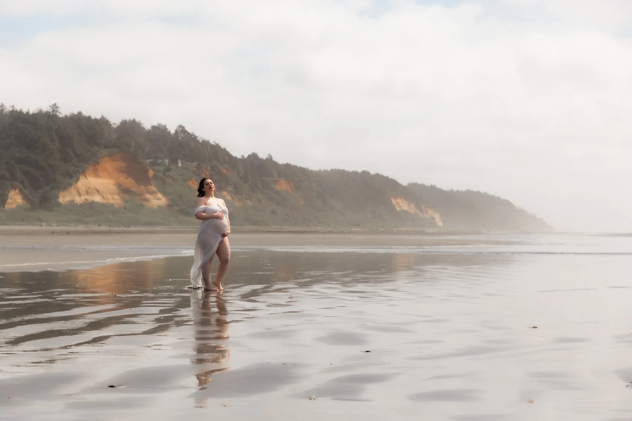A pregnant woman in a flowing dress standing on the beach with waves at her feet, cliffs in the background, and a cloudy sky.