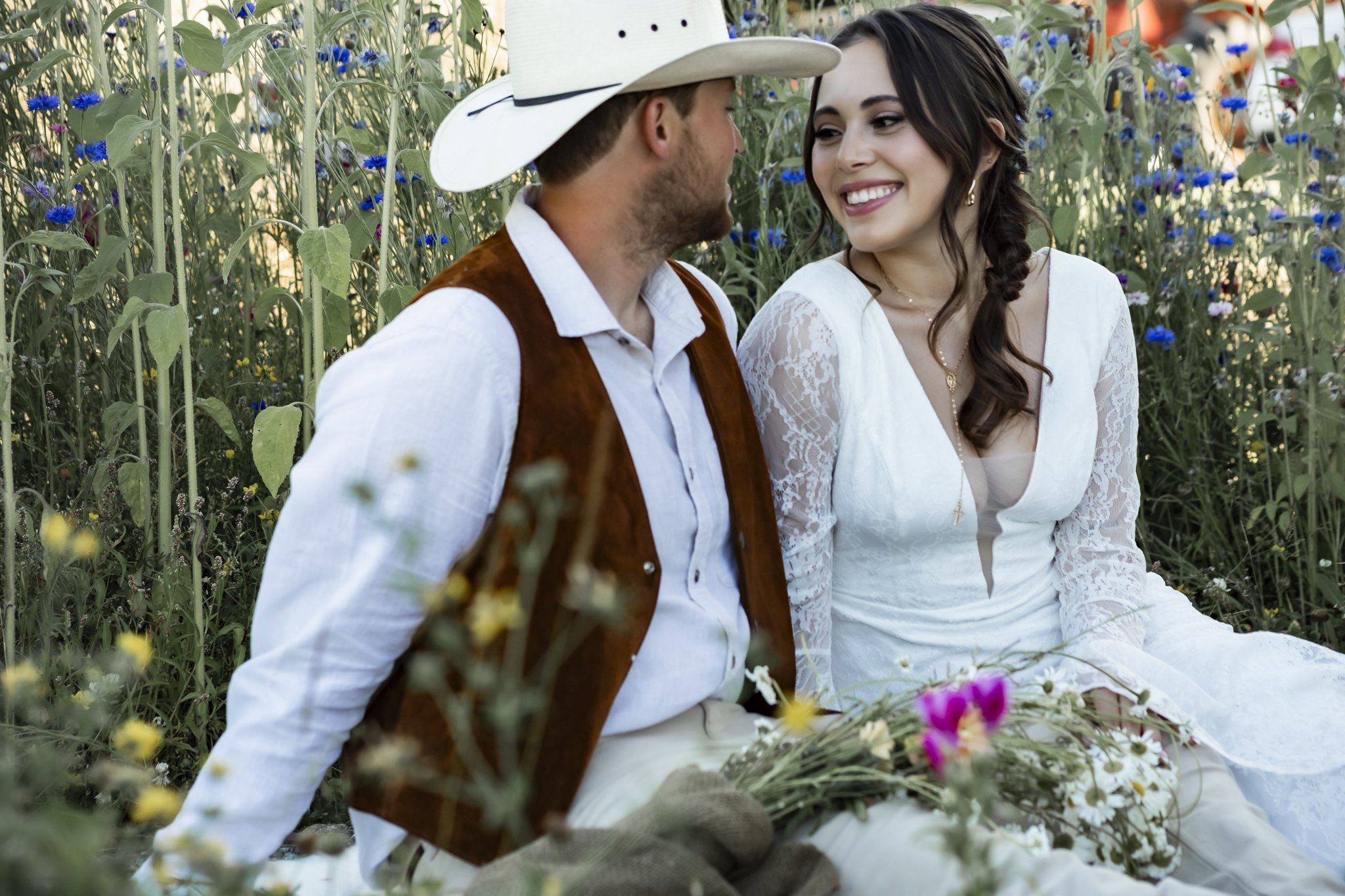 A man and woman sitting close together in a flower garden, smiling at each other. The woman is wearing a white lace dress and the man has a cowboy hat and brown vest. There are colorful flowers around them.