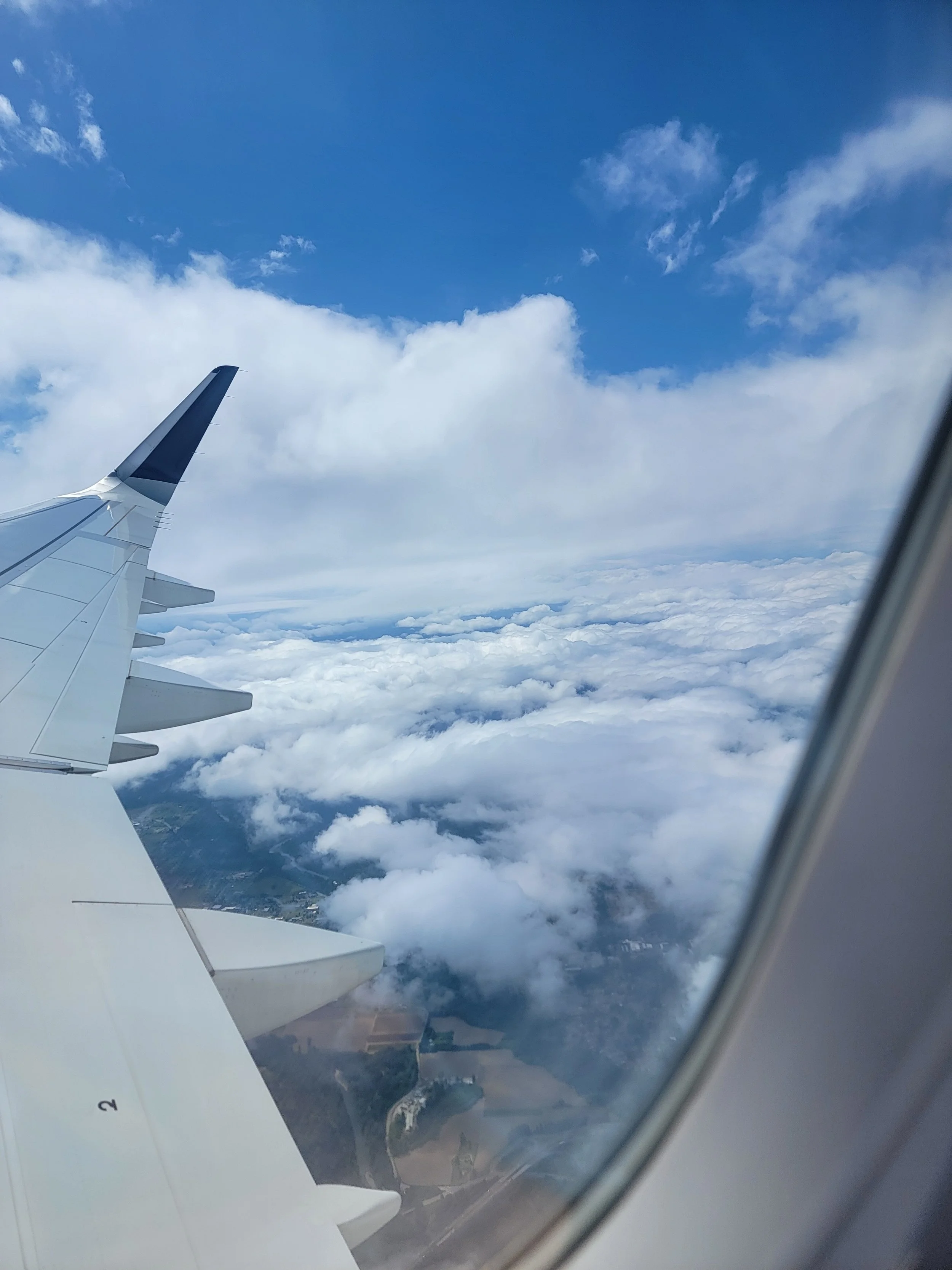 Courtney of Indulgence's view from an airplane window showing part of the airplane wing and a sky filled with clouds, with a glimpse of the landscape below.