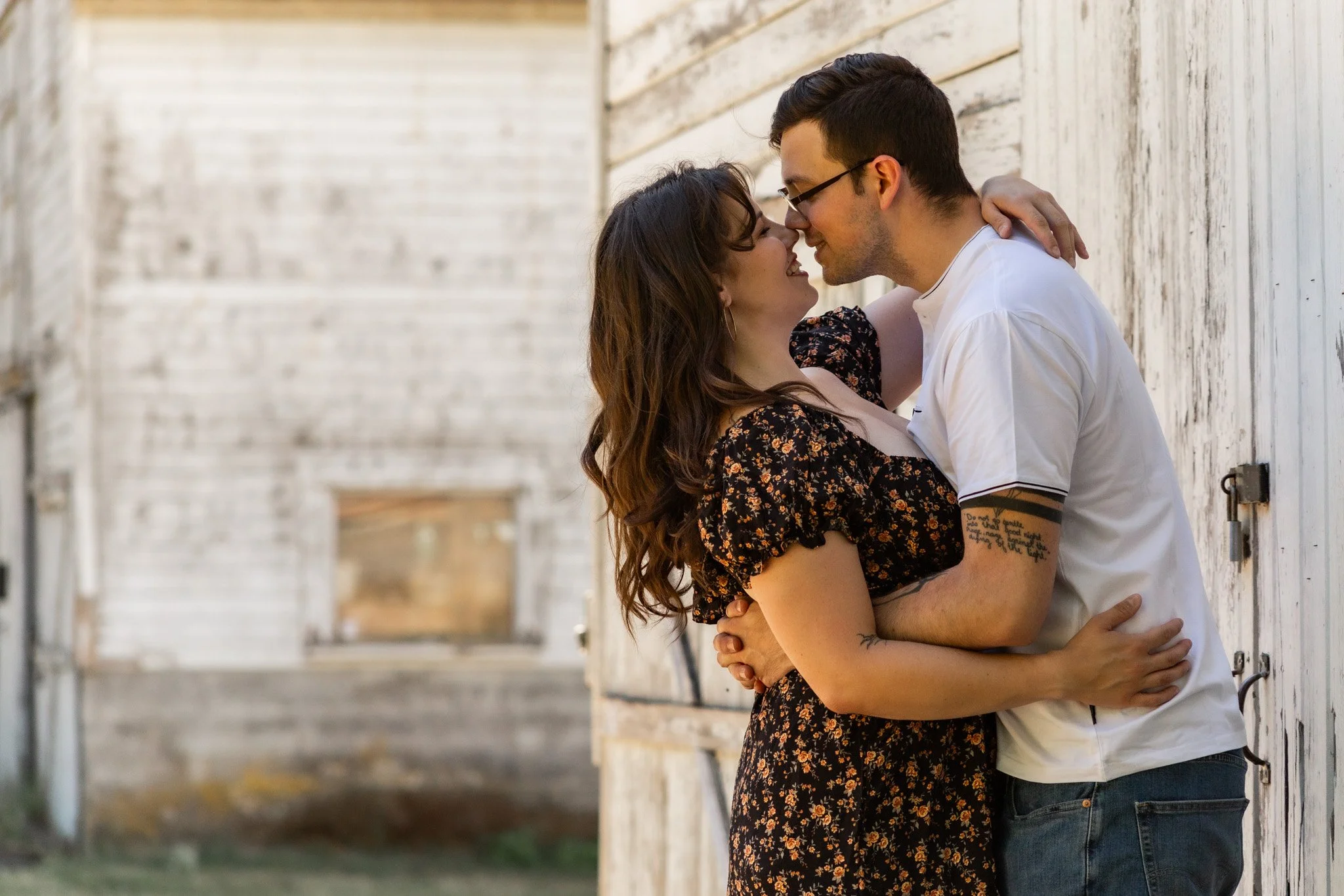 A young couple standing close to each other, touching noses, and smiling while embracing against a white wooden barn wall.