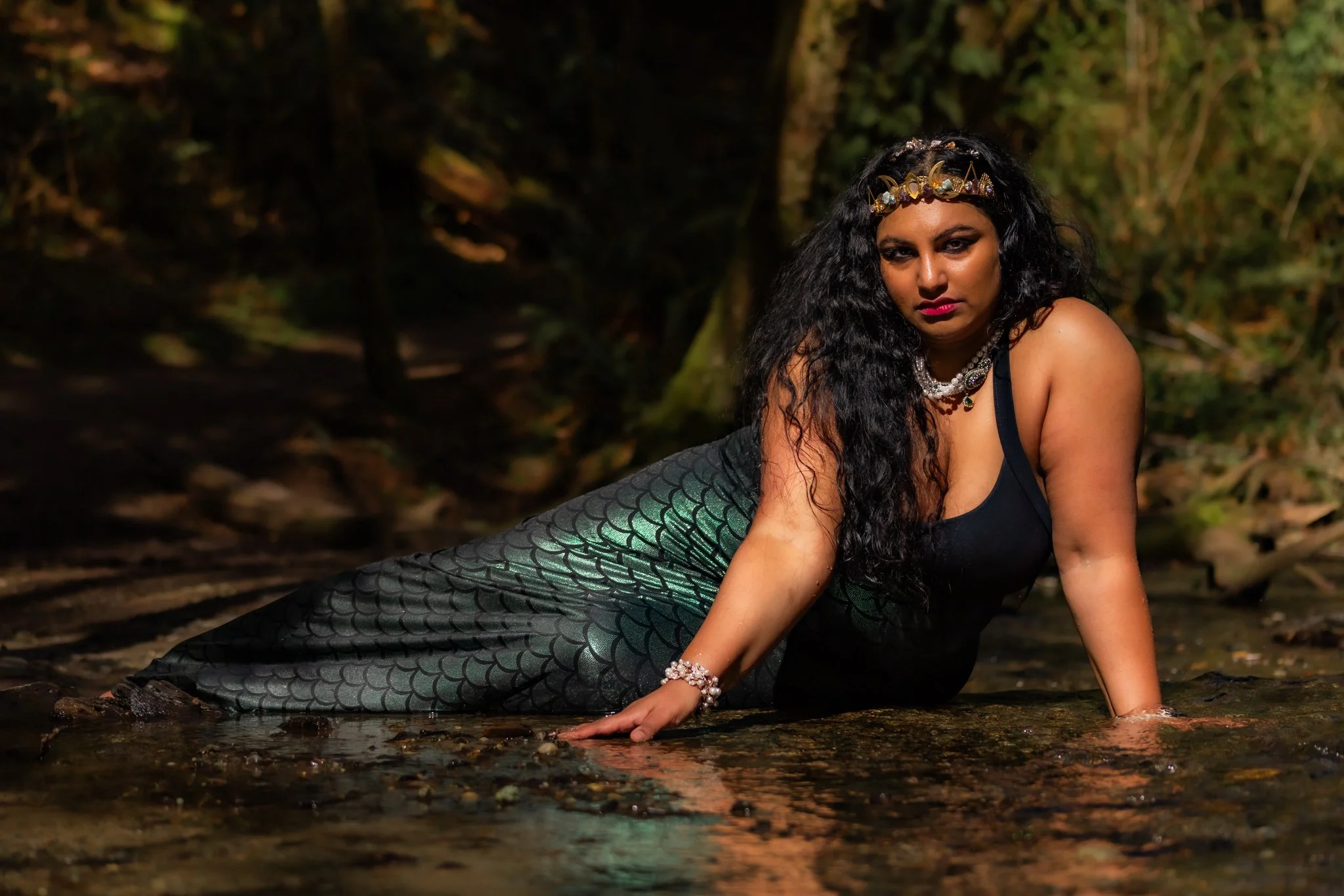 A woman dressed as a mermaid with long curly black hair, wearing a pearl and gemstone crown and necklace, lying on the ground in a shallow forest stream, with a dark forest background.