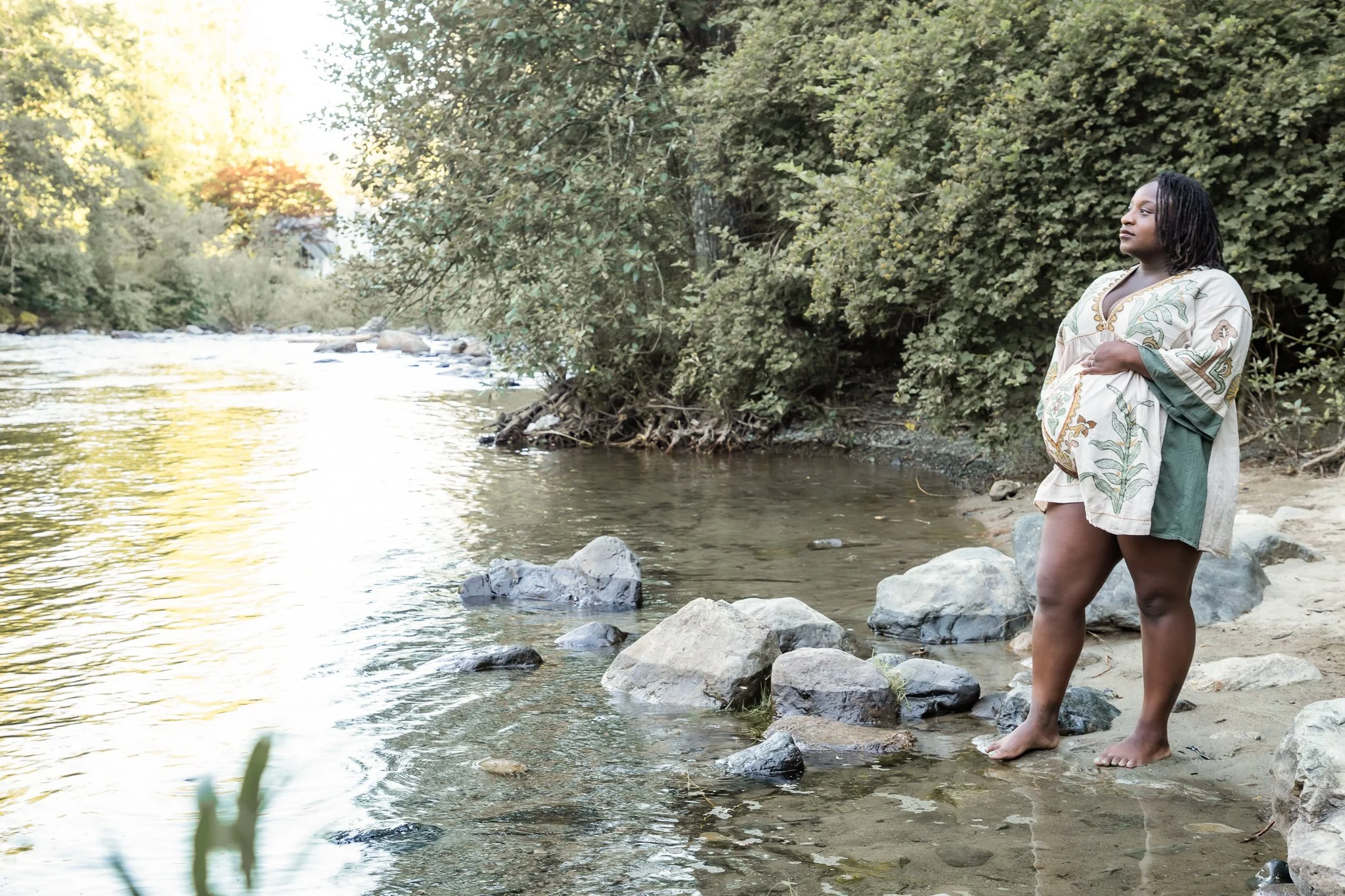 A woman standing barefoot on rocks at the edge of a river, surrounded by lush green trees, during sunset.