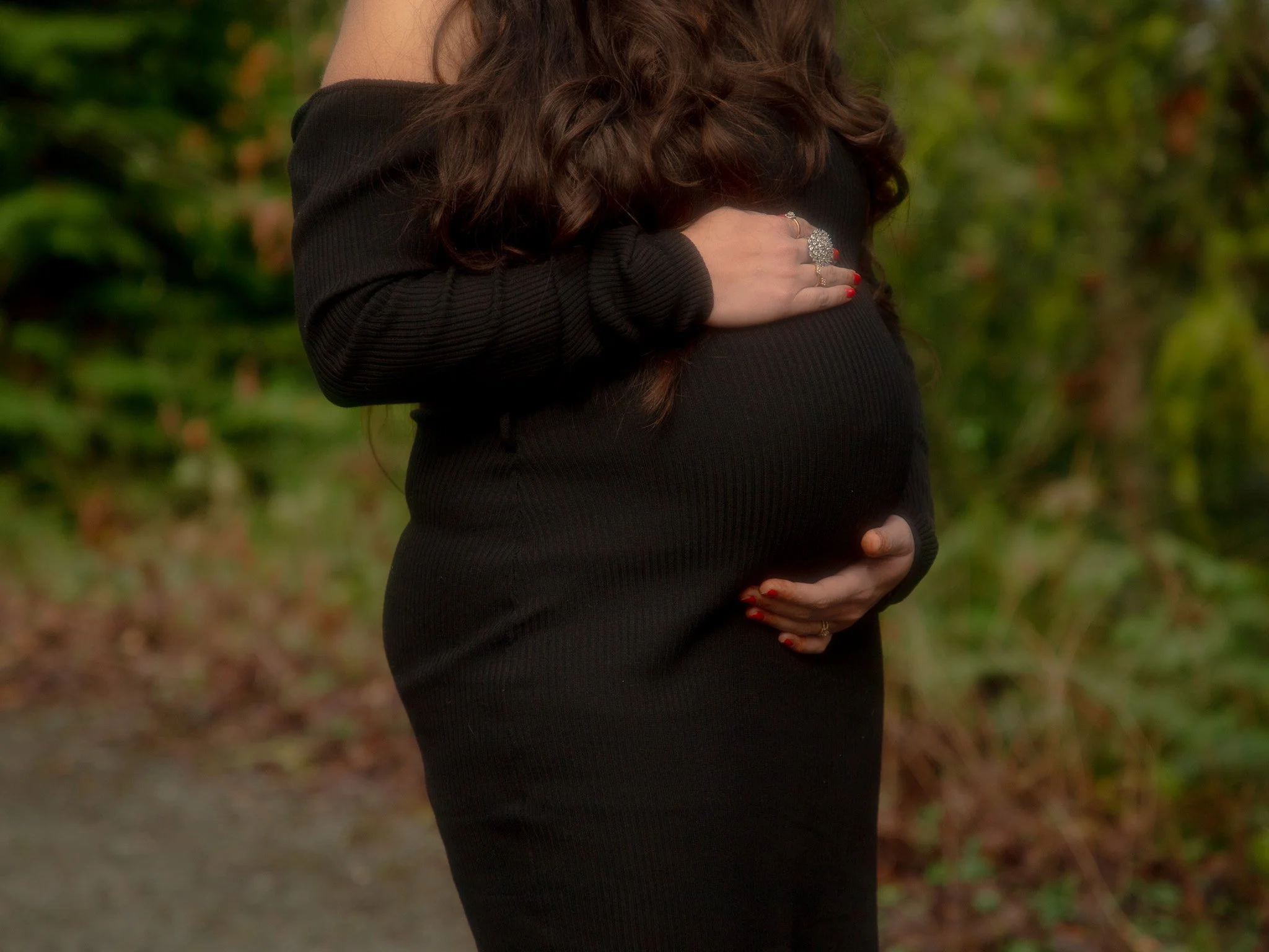 A woman with long, curly brown hair wearing a black long-sleeved top, holding her pregnant belly with both hands. She has painted red nails and is wearing a sparkling ring on her left hand. The background consists of green blurred foliage, indicating an outdoor setting.