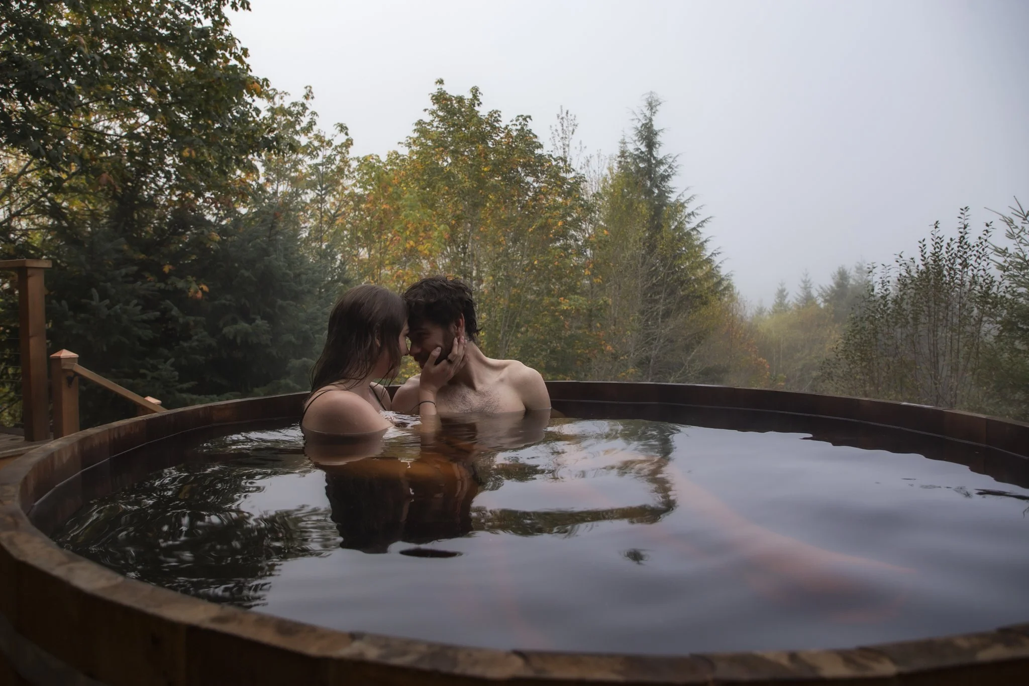 A couple sharing an intimate moment in an outdoor hot tub on a foggy day, surrounded by trees with autumn foliage.