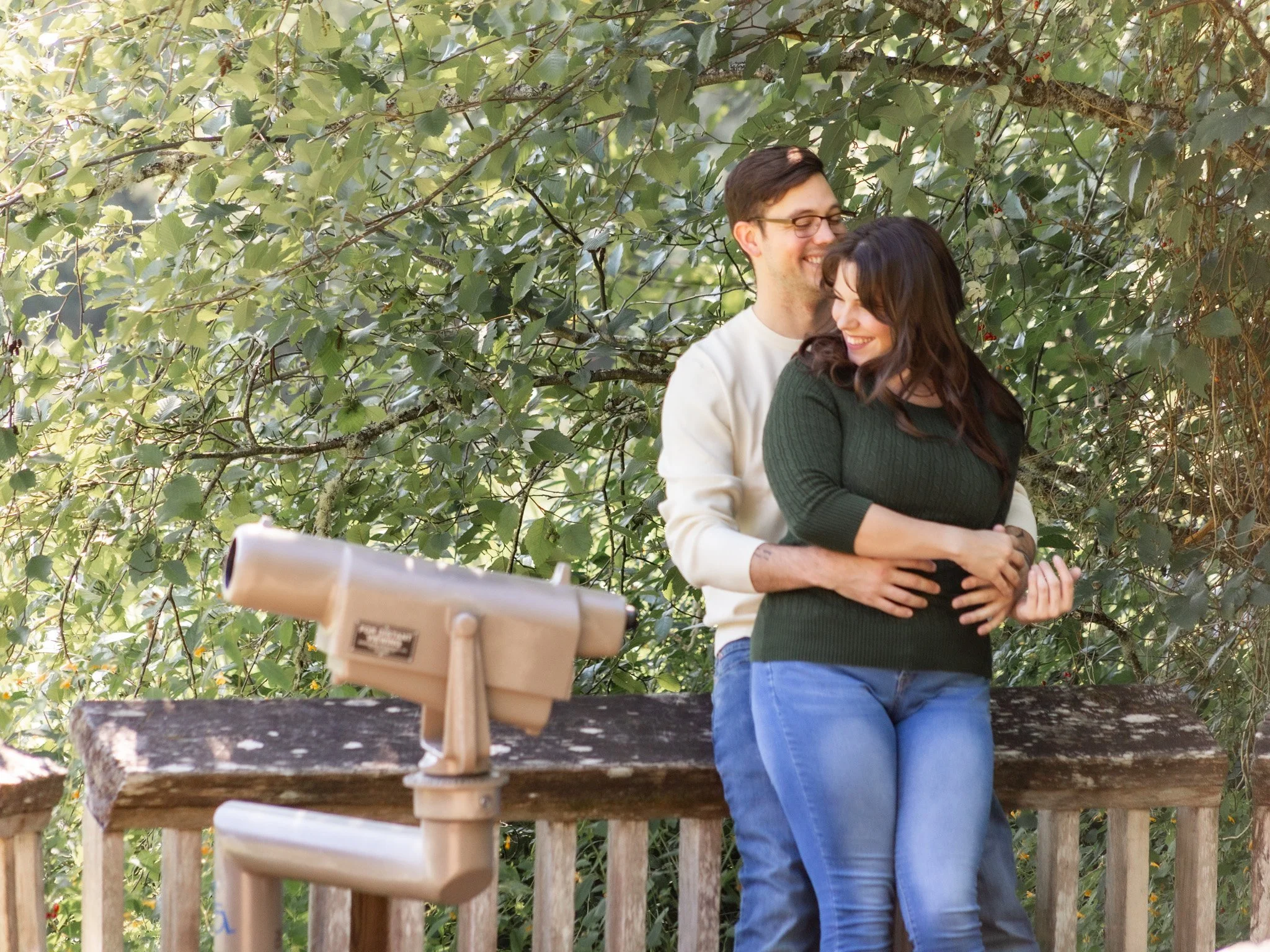 A smiling man and woman hugging on a wooden deck outdoors, surrounded by green foliage, with a video camera on a tripod in the foreground.