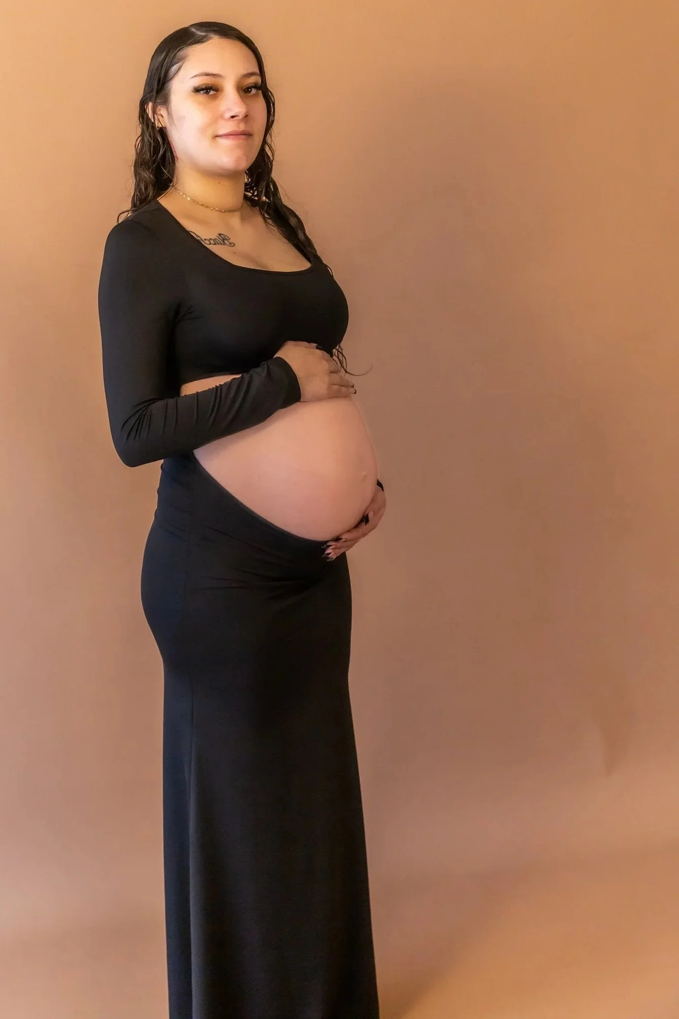 A pregnant woman with long dark hair, wearing a black long-sleeve top and black pants, stands profile against a plain beige wall. She is gently holding her baby bump with one hand and has a calm expression.