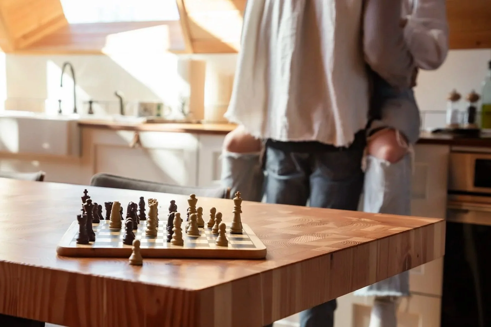 A chessboard with chess pieces set up on a wooden table in a brightly lit kitchen, with a person in casual clothing standing nearby.