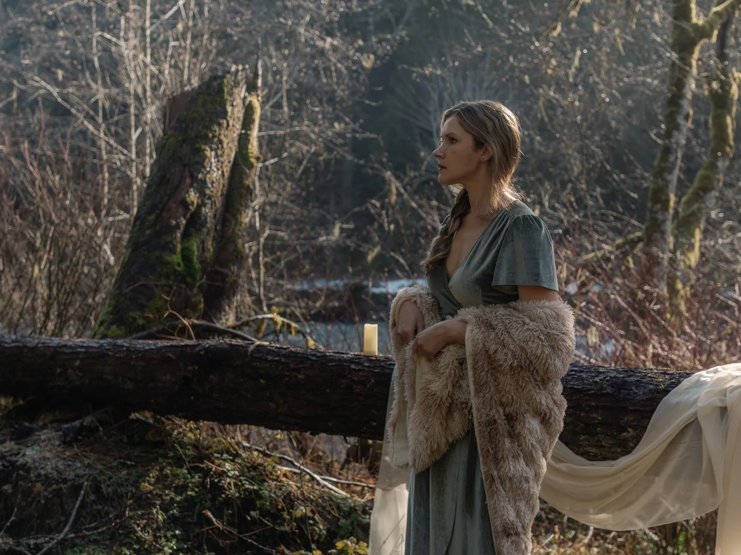 A woman with long hair styled in a braid standing outdoors among trees during daylight. She is wearing a green dress, a fur wrap around her shoulders, and a flowing cream fabric draped behind her. There is a small candle near her on a tree branch.