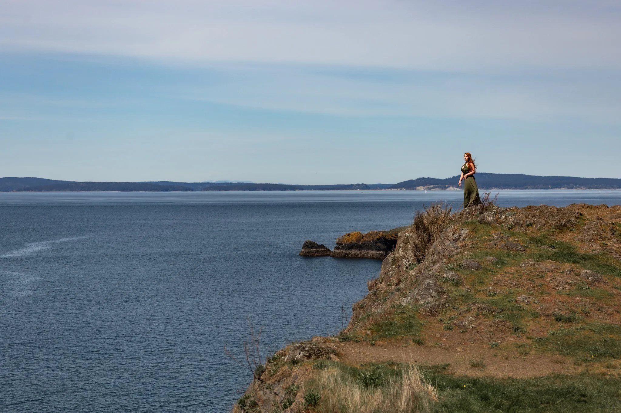 A woman standing on a rocky cliff overlooking a large body of water with distant hills under a partly cloudy sky.