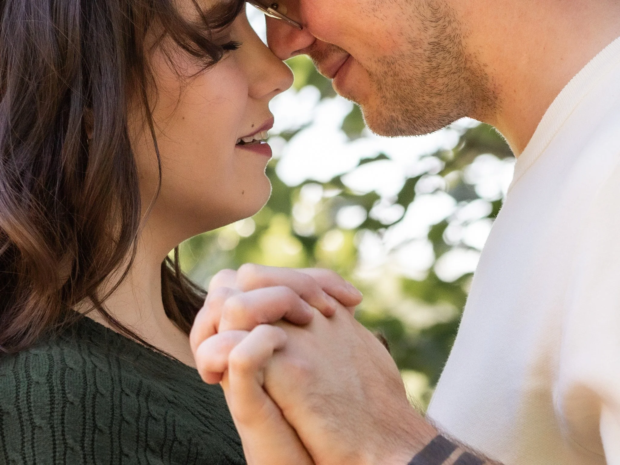 A young woman and a man with glasses touching foreheads and holding hands, outdoors with greenery in the background.