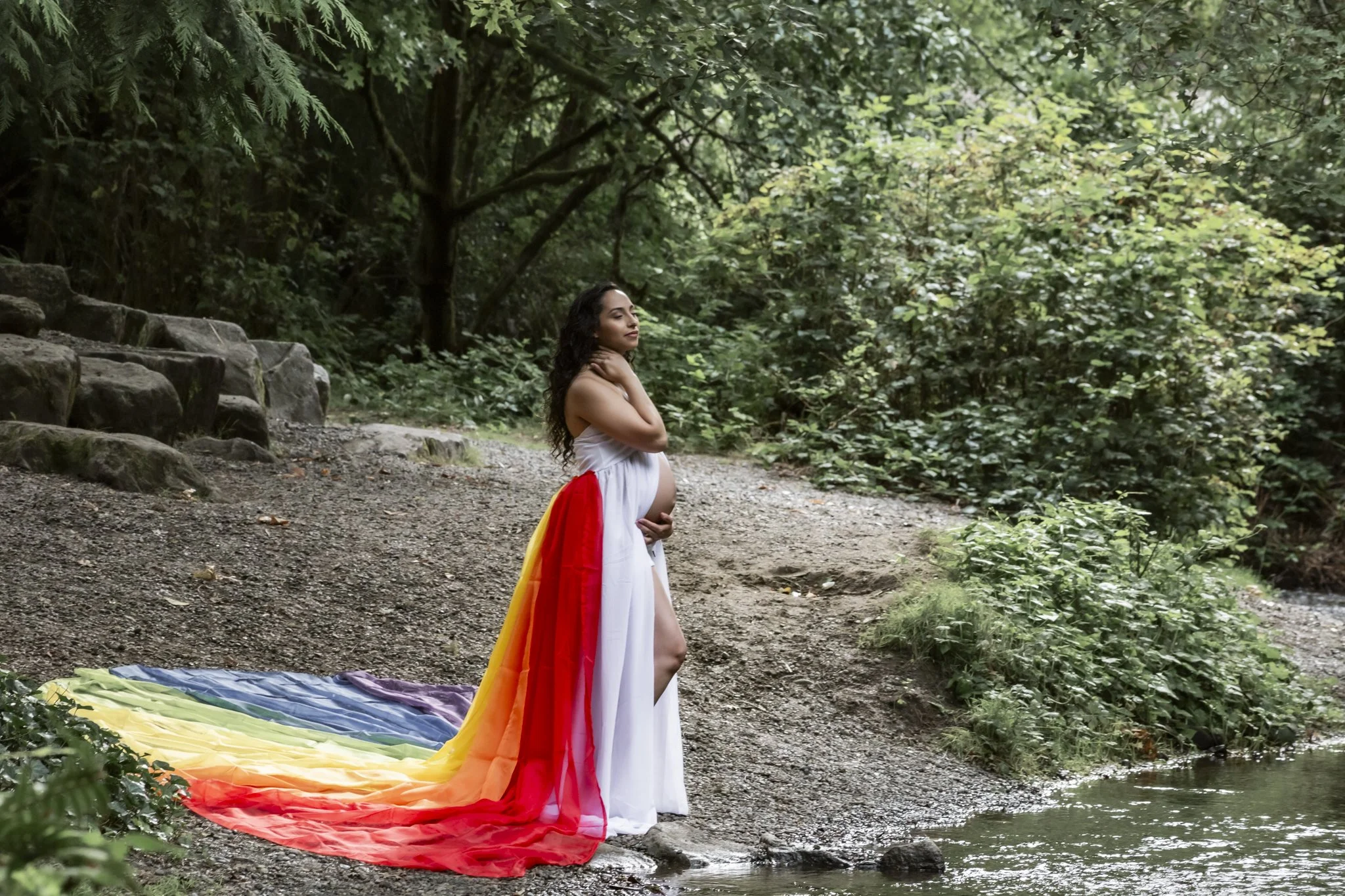 A pregnant woman dressed in a white gown with a rainbow-colored train stands near a stream in a forest, with her hand on her neck, looking peacefully into the distance.