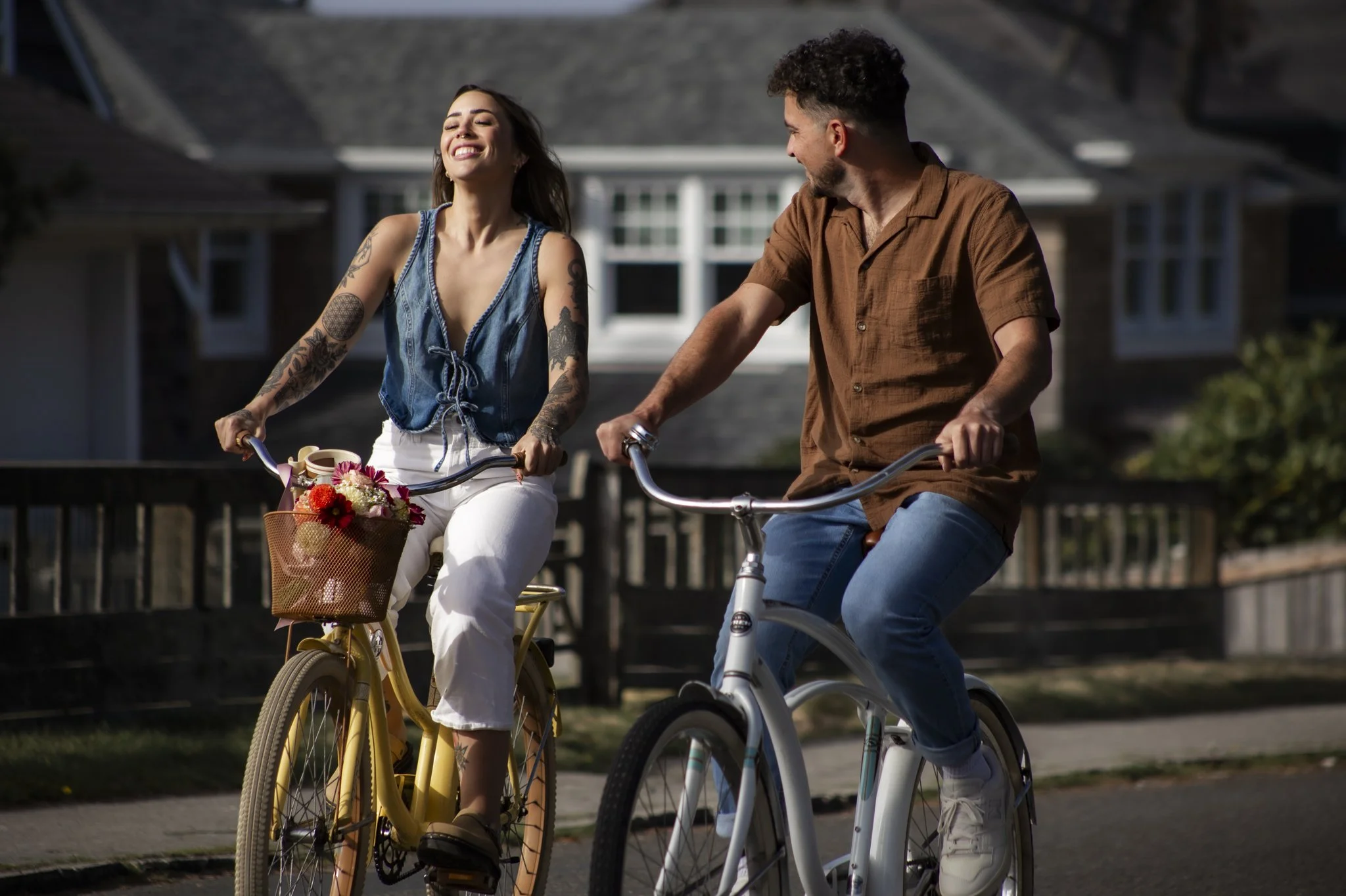 A woman with tattoos on her arms and long hair smiling happily while riding a yellow bicycle with a basket of flowers, accompanied by a man with curly hair and a beard wearing a brown shirt, riding a white bicycle.