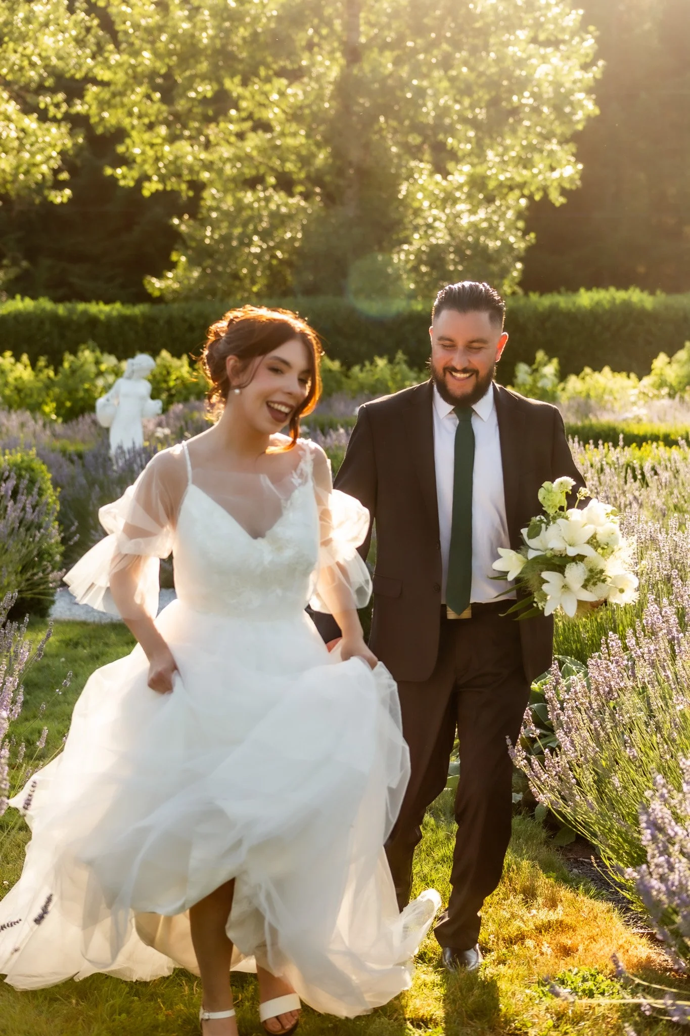 A bride and groom walking in a garden during a wedding celebration. The bride is wearing a white wedding dress and the groom is in a dark suit with a green tie. They are smiling and enjoying the moment in the sunlight surrounded by greenery and purple flowers.
