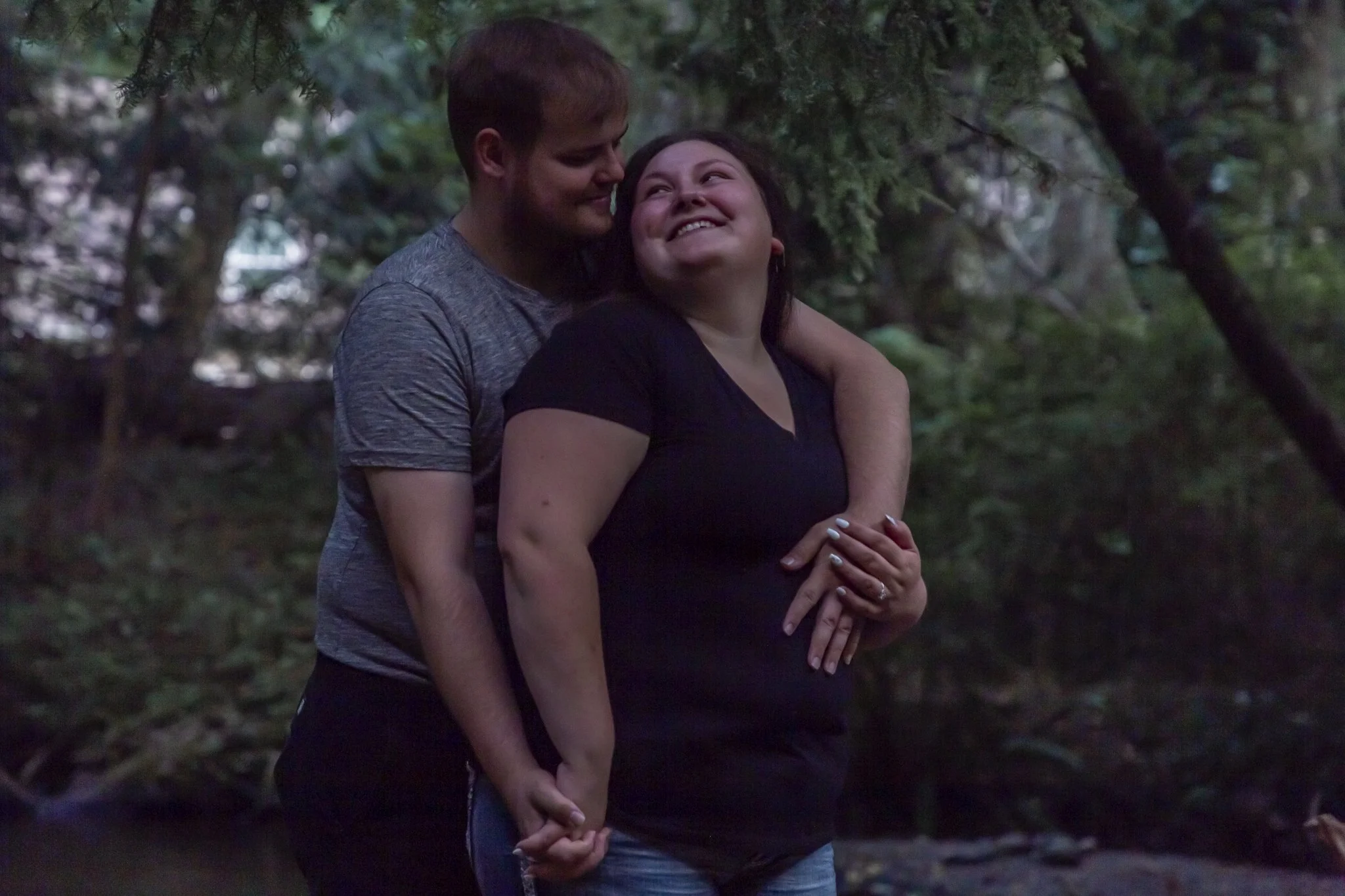 A couple embracing outdoors in a wooded area, with one person smiling and the other looking at them lovingly.