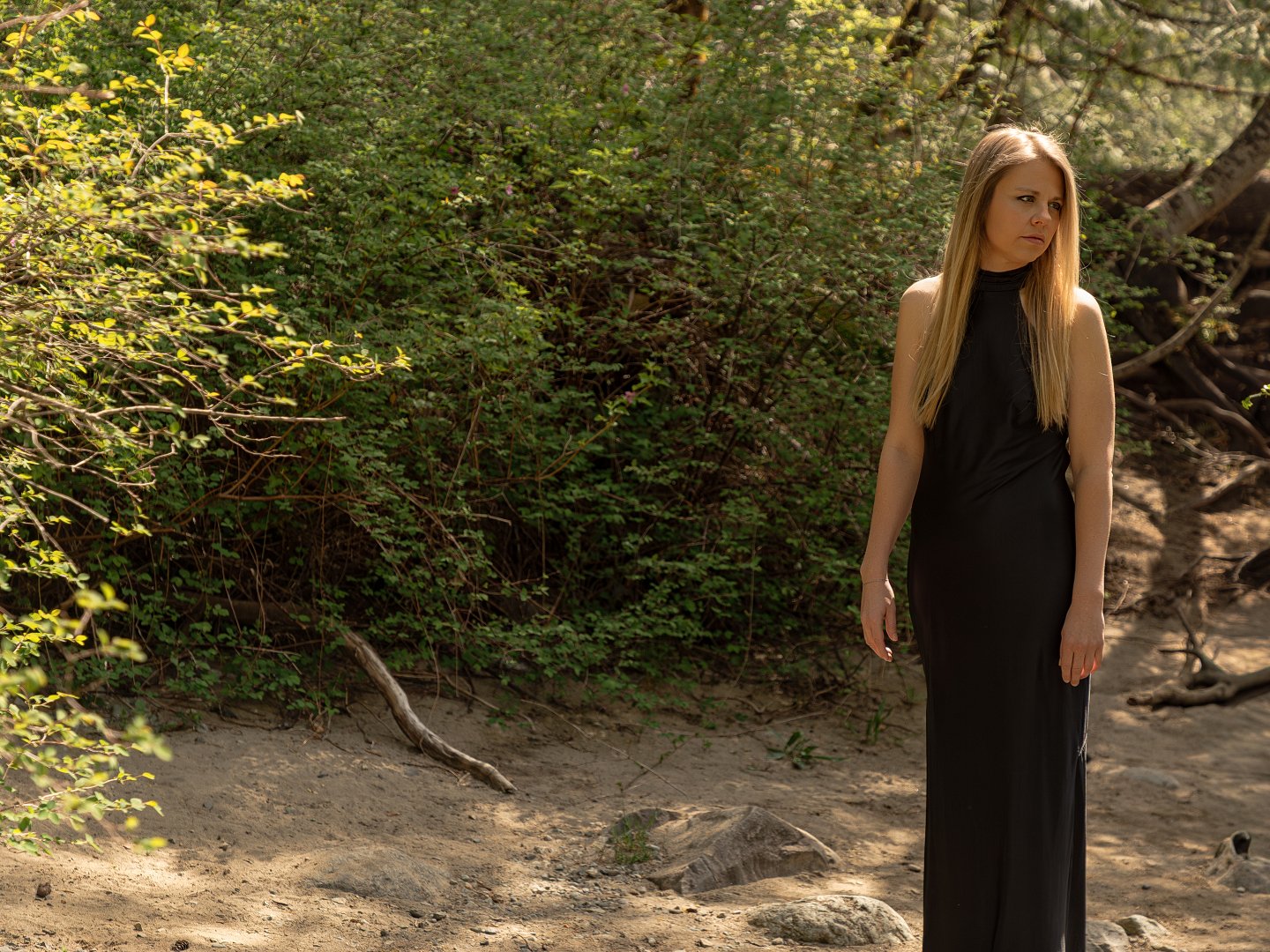 A woman with long blonde hair wearing a sleeveless black dress standing outdoors on a dirt path near green bushes and trees.