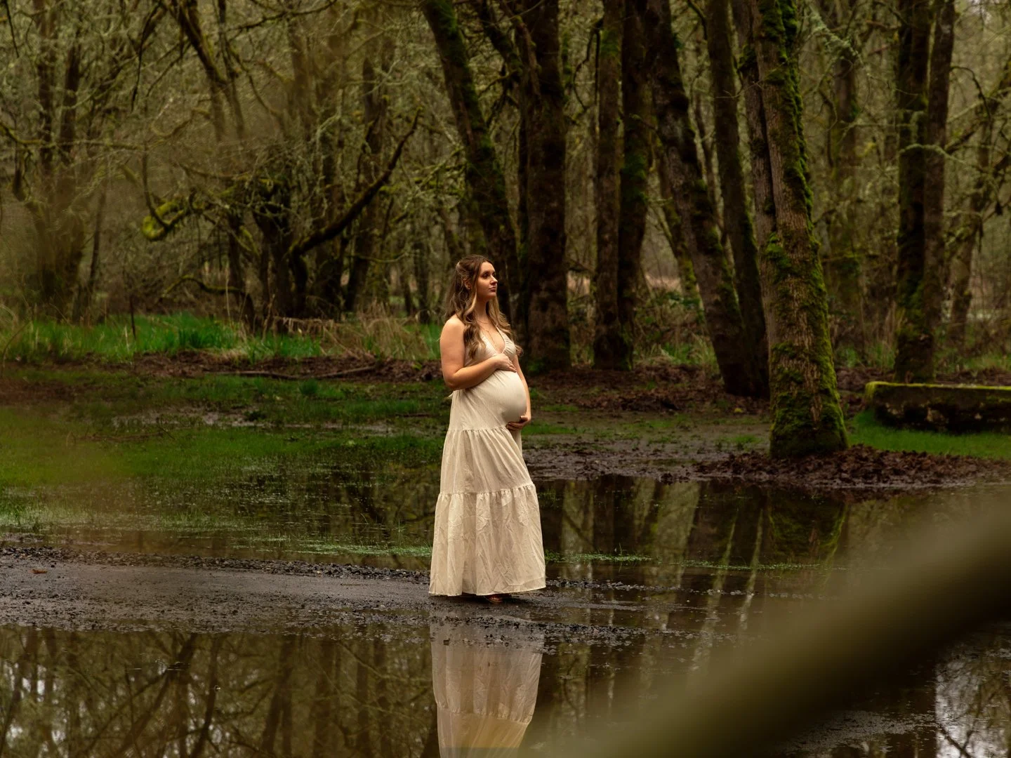 A pregnant woman in a long beige dress stands in a forest near a small body of water, holding her baby bump and looking peaceful.