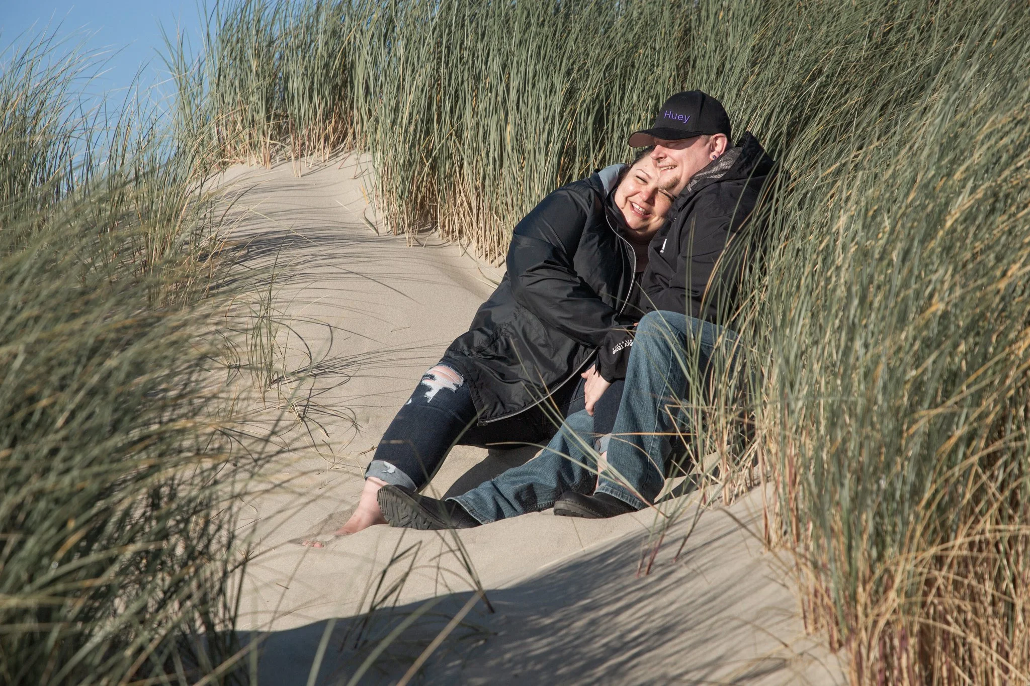 Two people sitting closely together on a sandy pathway among dune grass on a sunny day, smiling and enjoying each other's company.