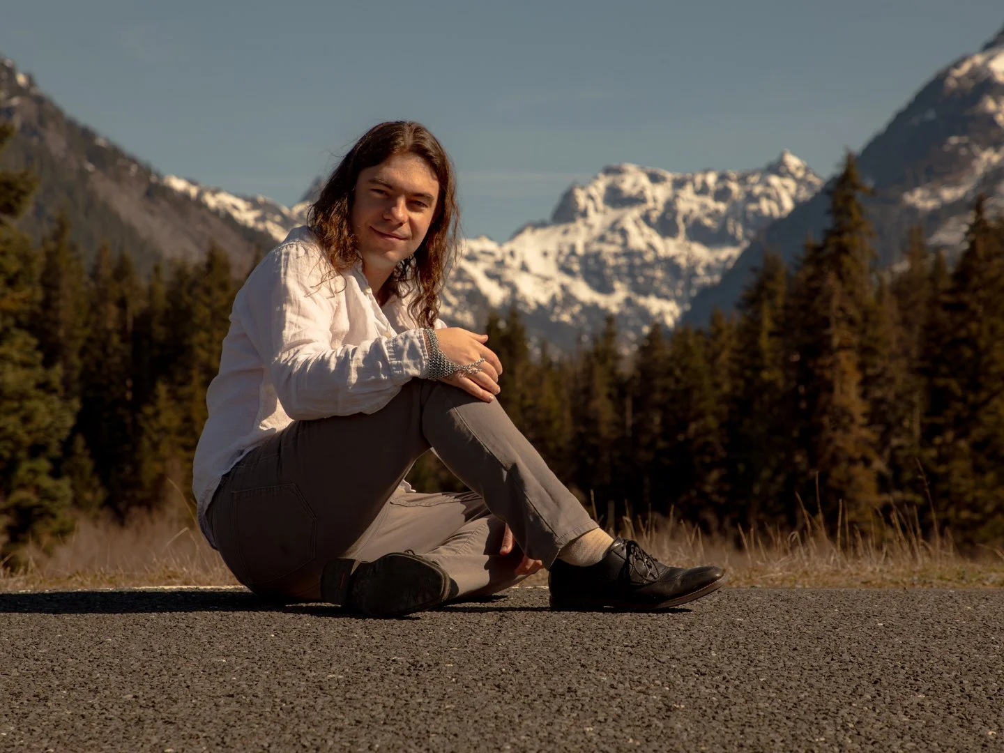 A person with long hair sitting on a road with a mountainous, snowy landscape and evergreen trees in the background.