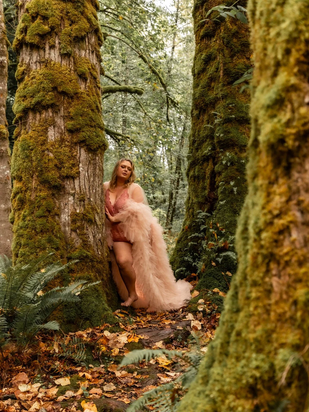 A woman in a glamorous peach-colored dress with long tulle train stands between two moss-covered trees in a forest, surrounded by fallen leaves and greenery.
