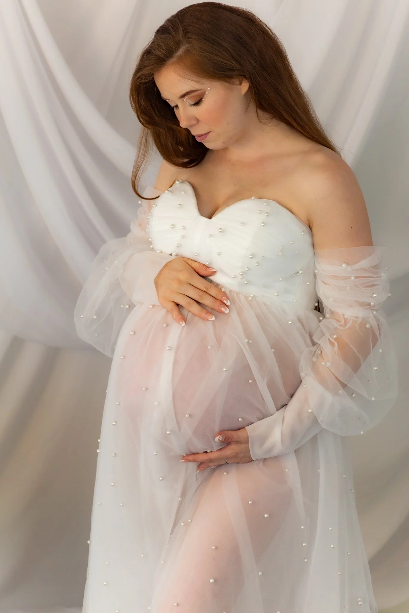 A woman with red hair is wearing a sheer, white dress with pearl embellishments, standing against a draped white background, looking down with her hand on her stomach.