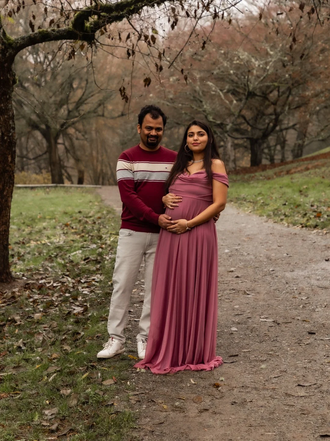 A pregnant woman in a pink dress and a man in a maroon and white striped sweater standing on a park path surrounded by trees and fallen leaves during autumn.
