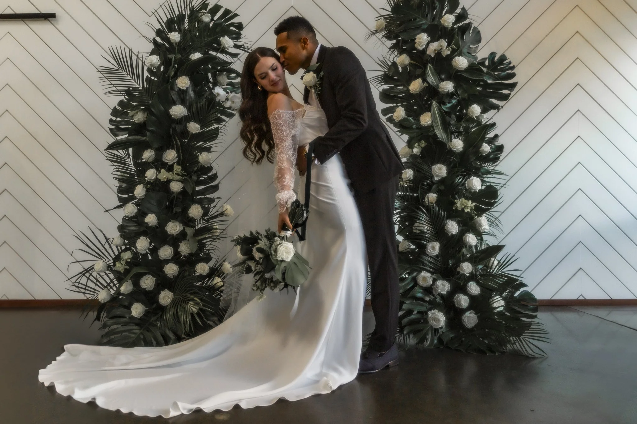 A bride and groom in wedding attire sharing a romantic moment in front of a floral backdrop with black leaves and white roses, on a white wall with a tilted panel detail.
