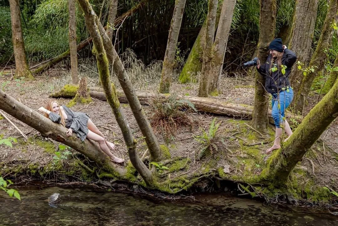 A woman is lying on a tree lying over a small stream in a forest, posing for a photo, while another woman takes her picture with a camera.