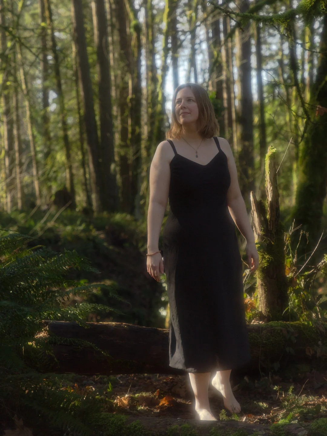 A woman with shoulder-length brown hair in a black dress standing barefoot in a forest with tall trees and green foliage.