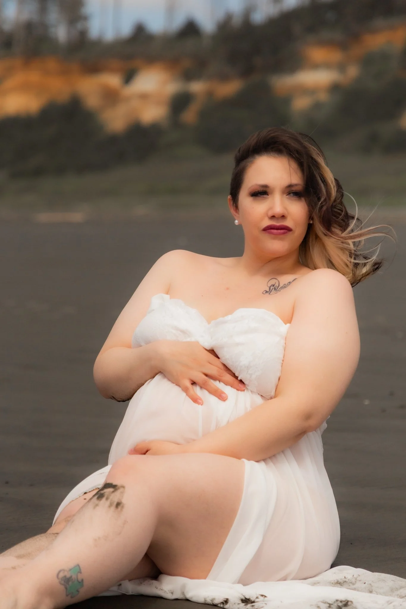 A woman with short hair and tattoos on her leg, wearing a white dress, sitting on a beach with a blurred background of cliffs.