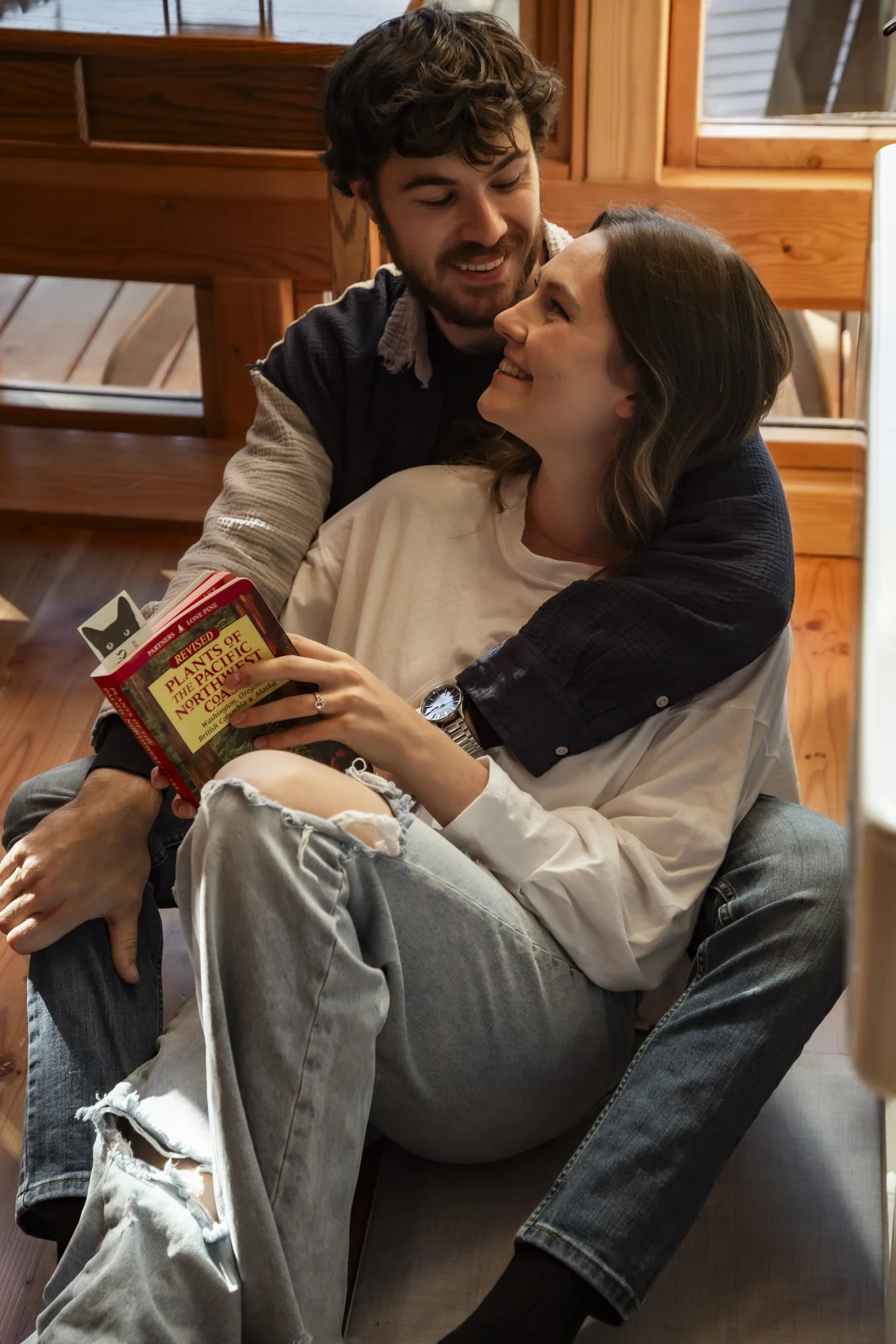 A couple sitting on the floor, smiling at each other. The woman is holding a book titled 'Plants of the Pacific Northwest' and has dark hair, wearing a white shirt and ripped jeans. The man has curly hair, a beard, and is wearing a dark jacket and black socks, embracing her from behind. They are in a cozy room with wooden walls and a window.