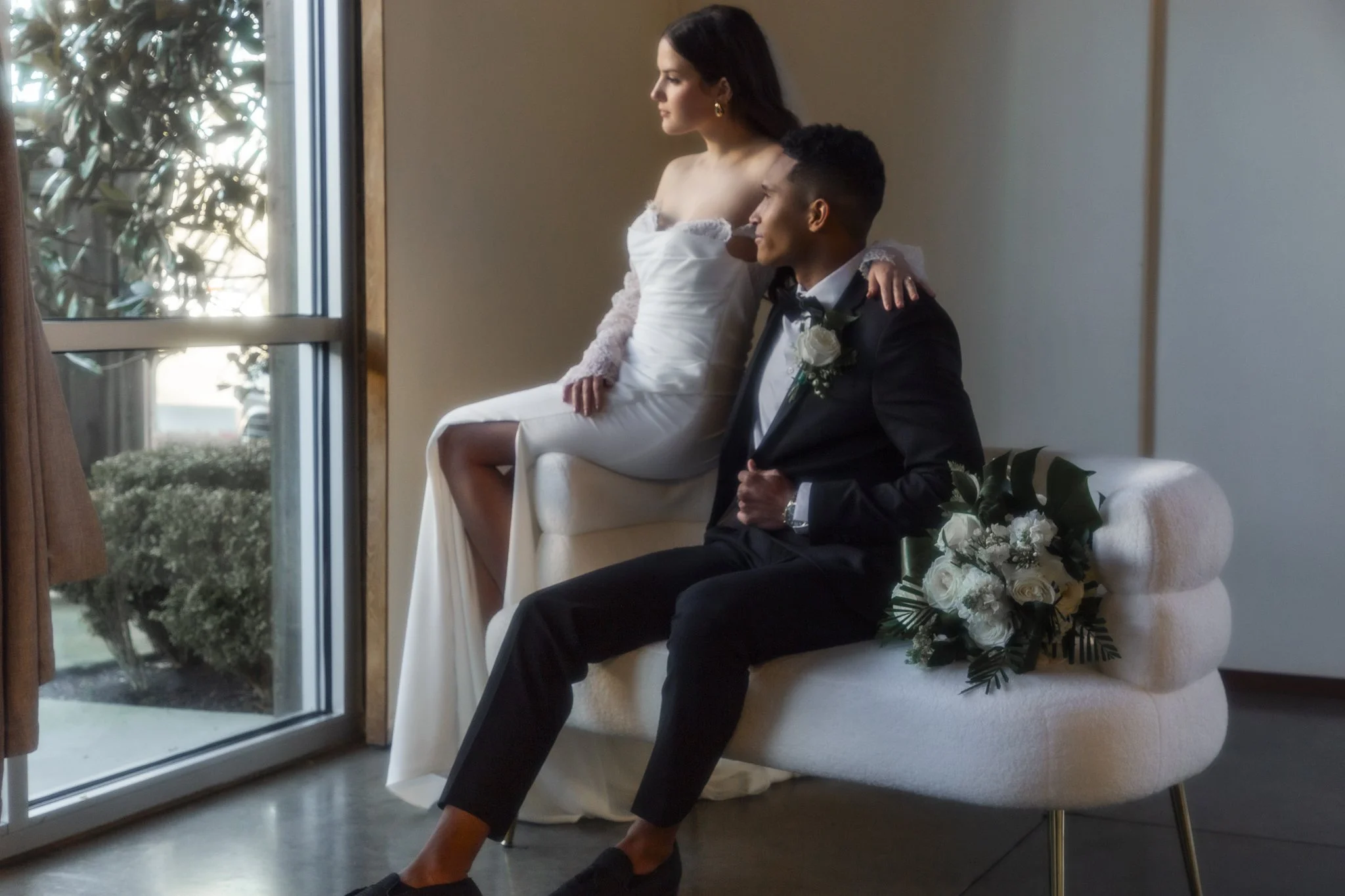 A woman in a white dress and a man in a black tuxedo sit side by side on a white upholstered bench, looking out a large window with sunlight streaming in. The woman has a bouquet of white roses resting on the bench beside her.