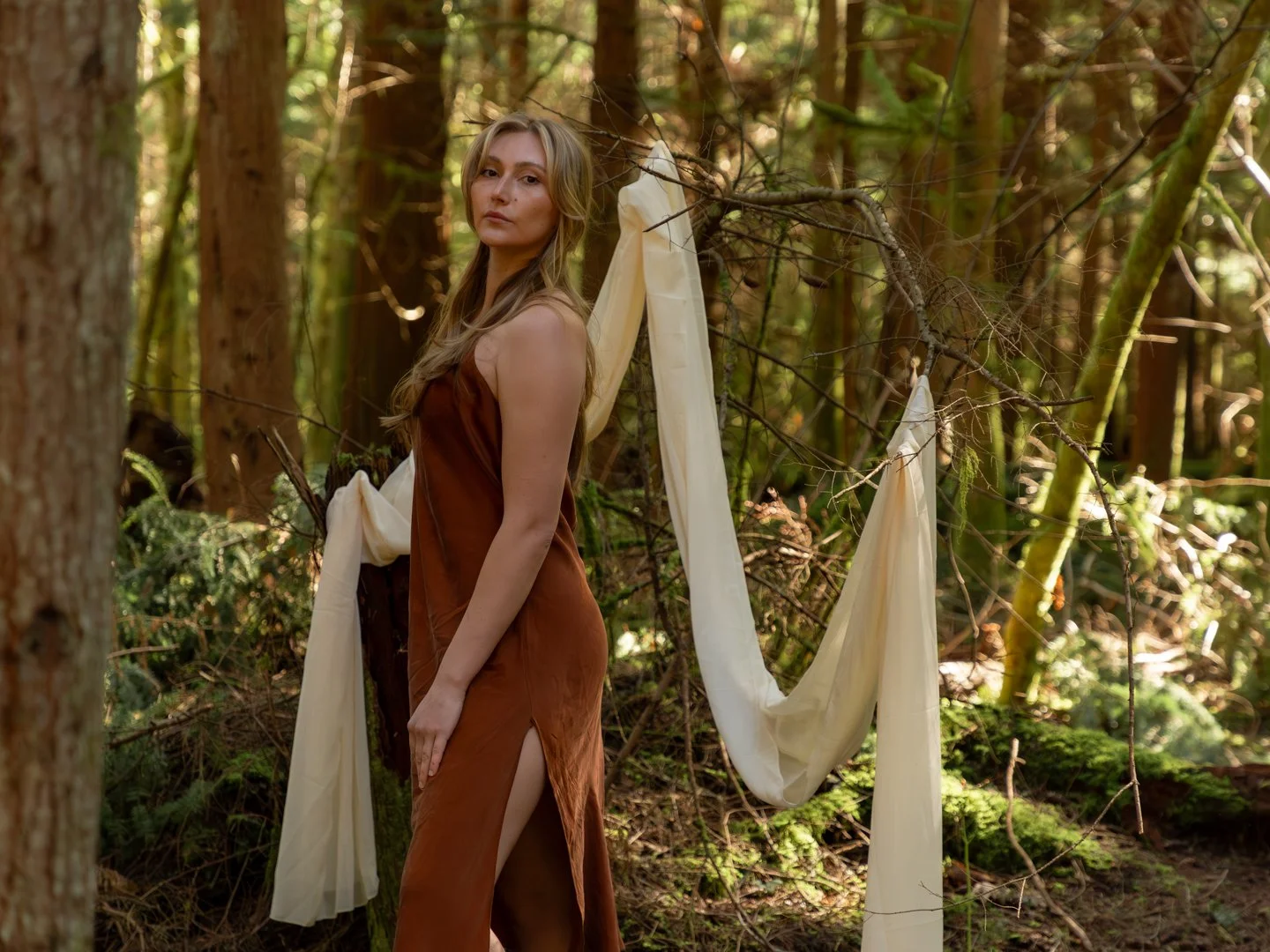 A young woman in a brown dress standing in a forest with beige fabric draped over a branch behind her.