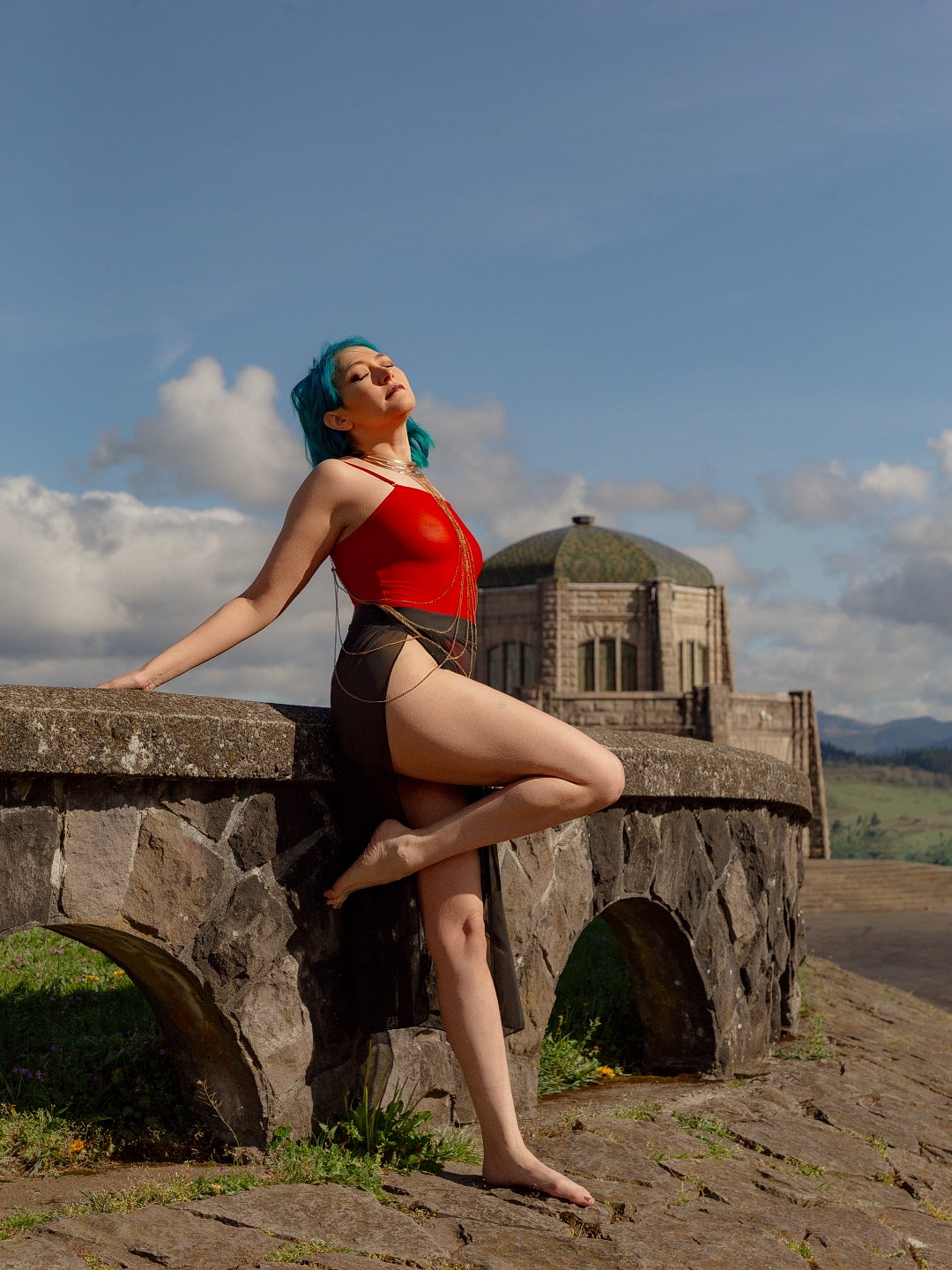 A woman with blue hair wearing a red top and black skirt posing outdoors on a stone bridge with a castle-like structure in the background under a partly cloudy sky.