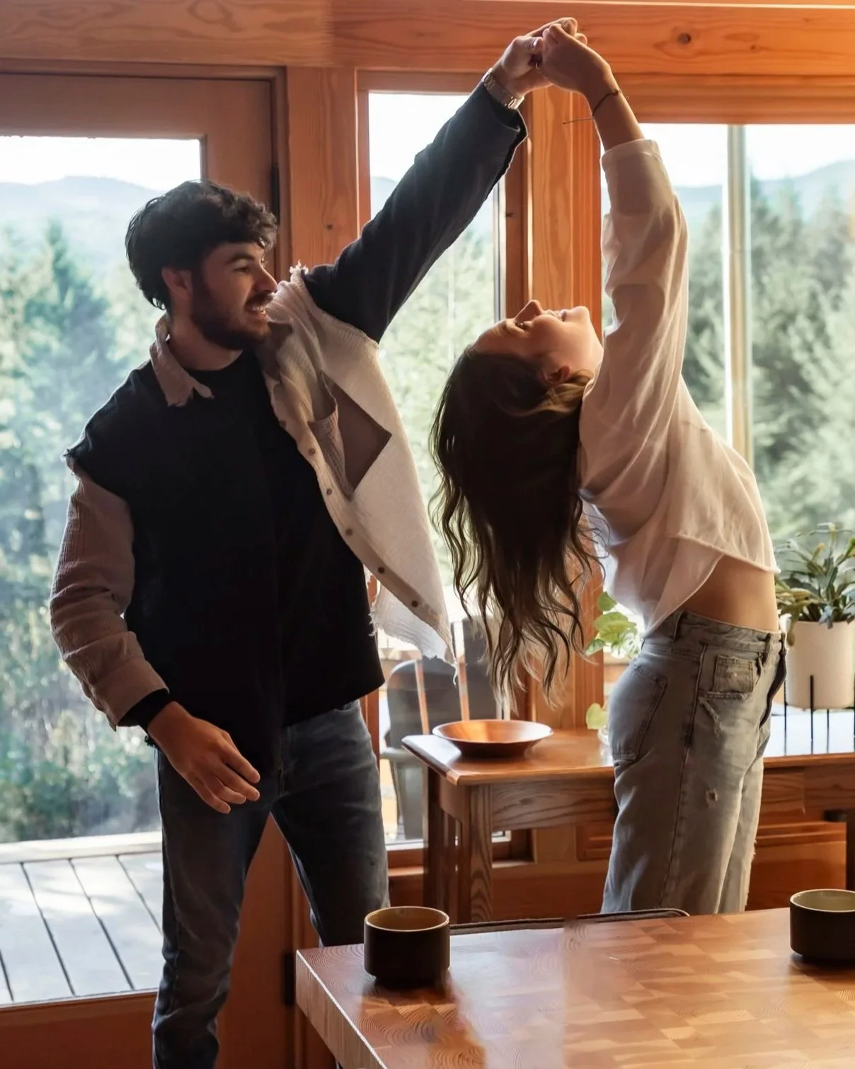 A man and woman dancing together inside a wooden house during daytime, with large windows showing a green outdoor landscape.