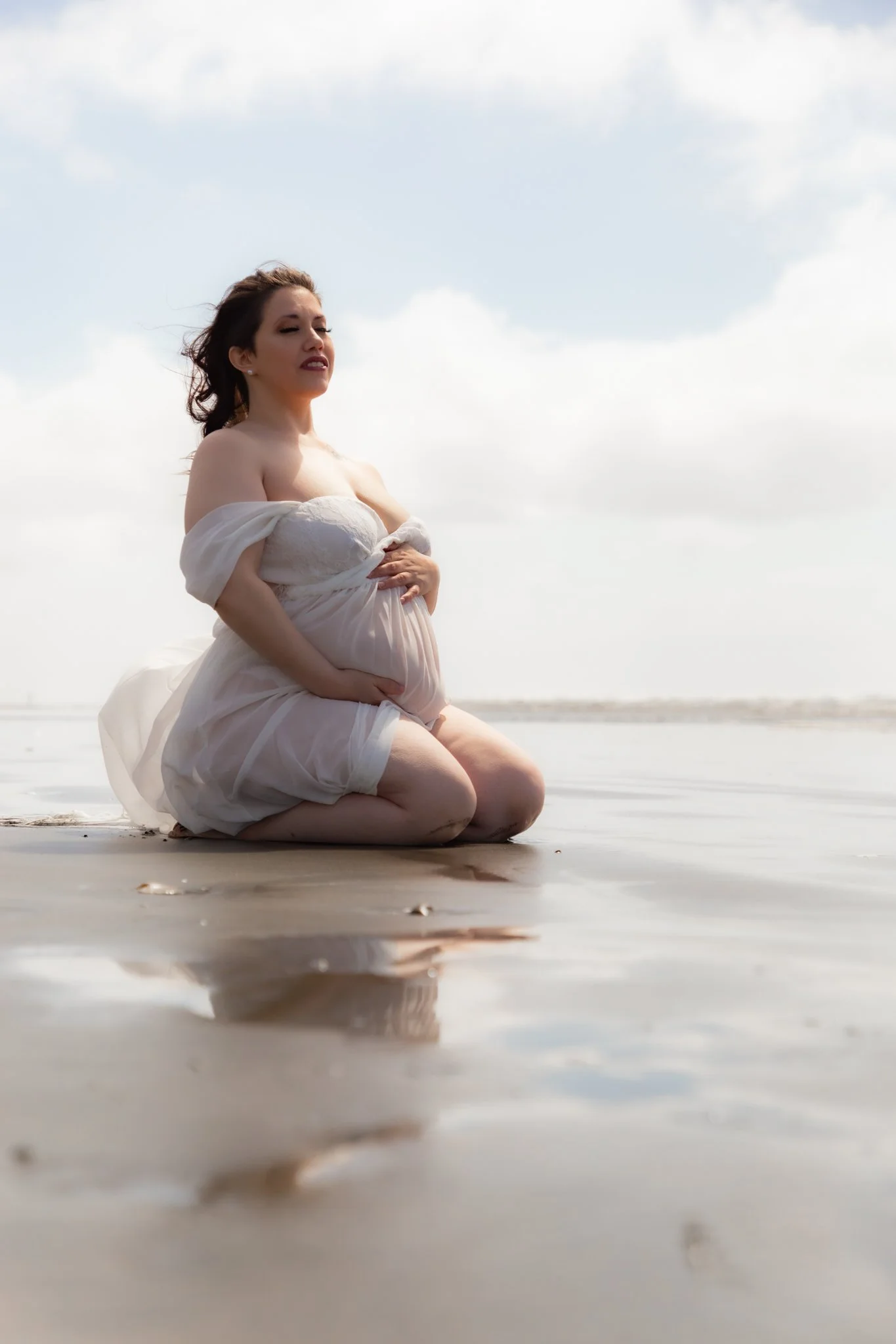 A pregnant woman kneeling on the beach in a white dress, with clouds in the sky and her reflection visible in the wet sand.