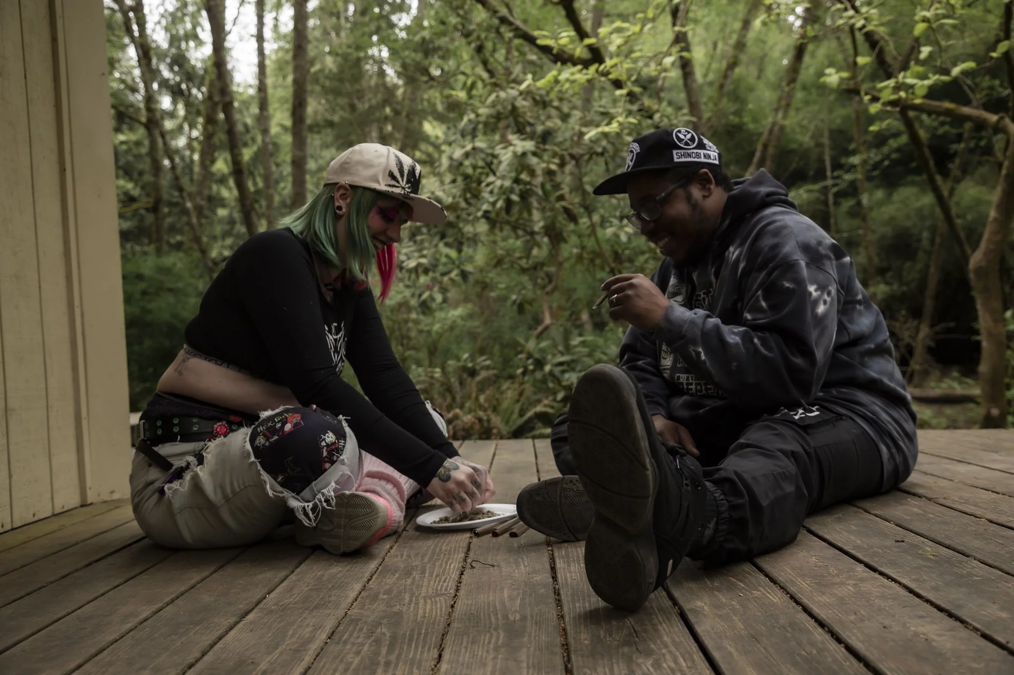Two people sitting cross-legged on a wooden porch in a forest, sharing a moment, with one person wearing a white cap with a black design and the other wearing a black hat with patches, both smiling.