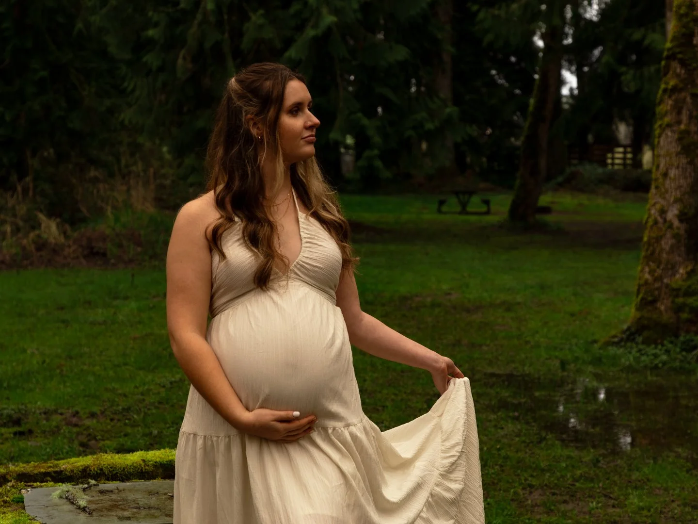 Pregnant woman in a flowing cream dress holding her belly and a part of her dress outdoors in a grassy, wooded area with trees and a rustic picnic table in the background.