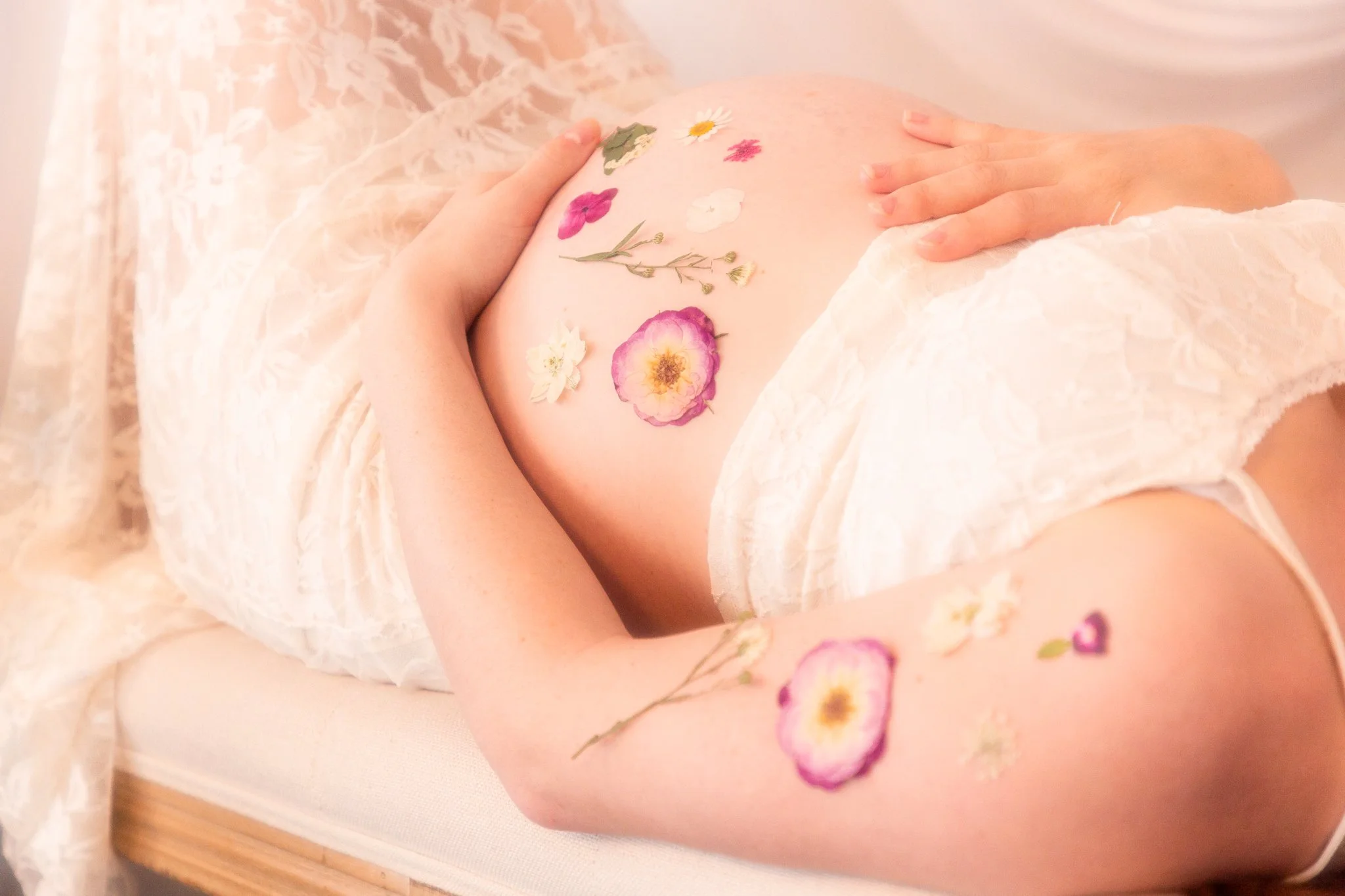A pregnant woman lying on a bed with her hand on her belly, which is decorated with various pressed flowers, including daisies and pink roses.
