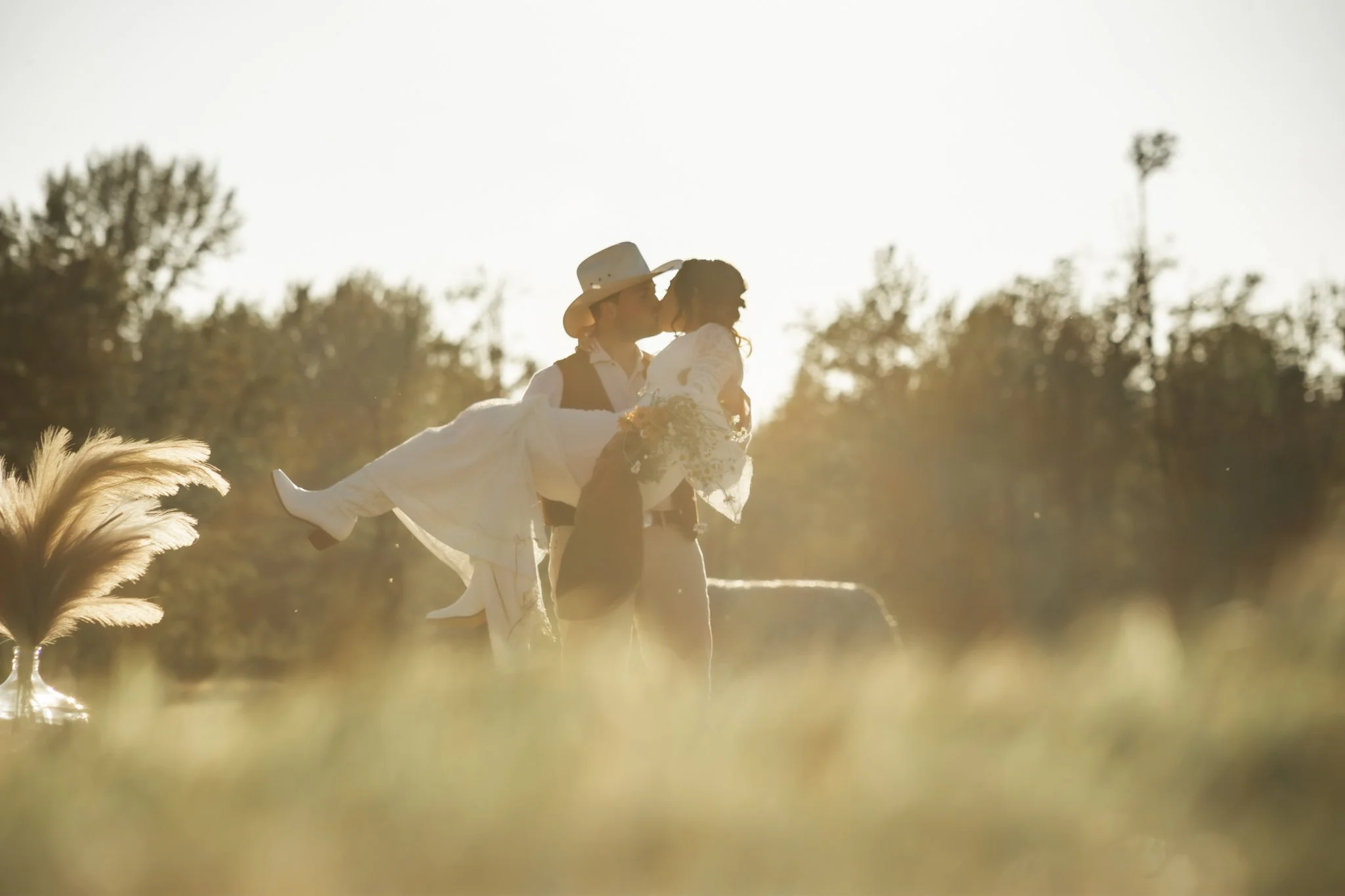 A couple dressed in wedding attire, with the groom wearing a cowboy hat, sharing a kiss outdoors during sunset, with trees in the background.