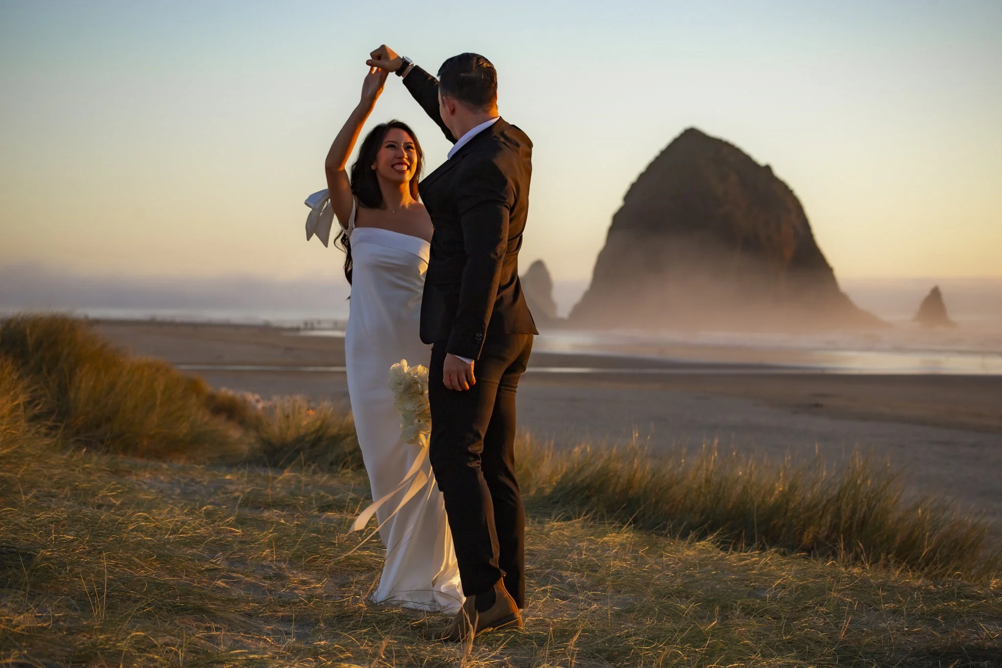 A bride and groom dancing on the beach at sunset with Haystack Rock in the background.